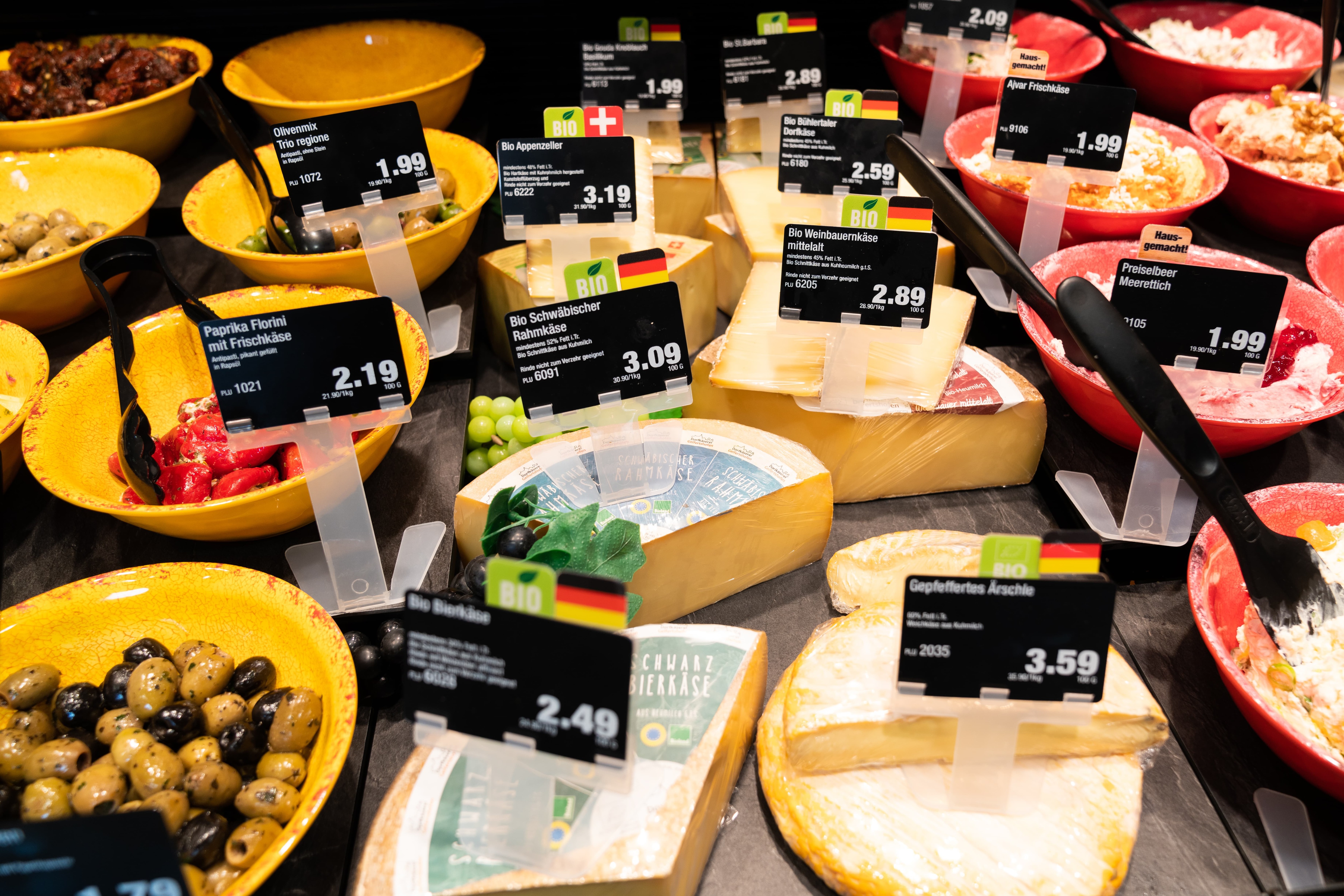 Verschiedene Käsesorten und Antipasti mit Preisschildern in einer Auslage.