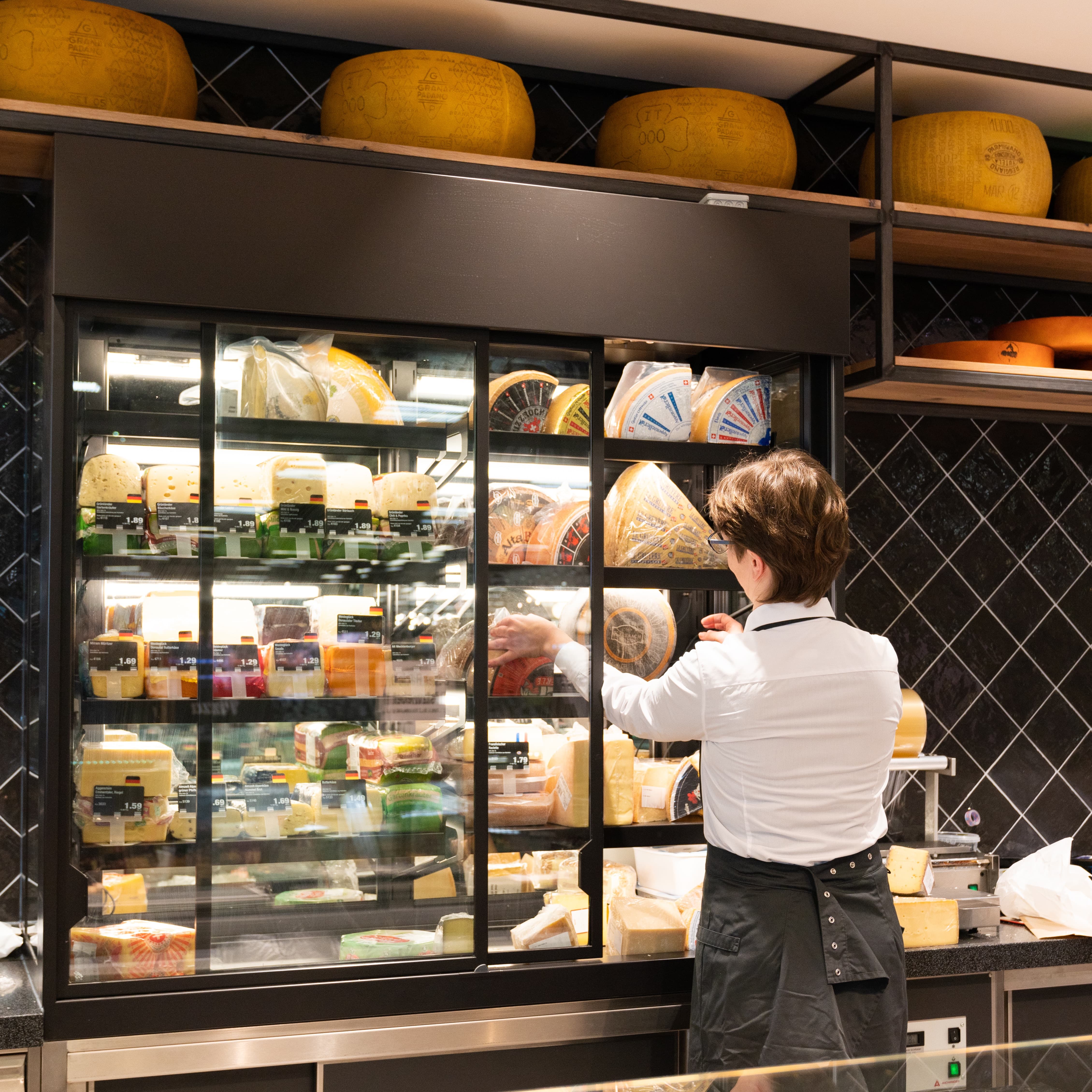 Eine Frau mit weißem Hemd und schwarzer Schürze greift in einen offenen Käseschrank.