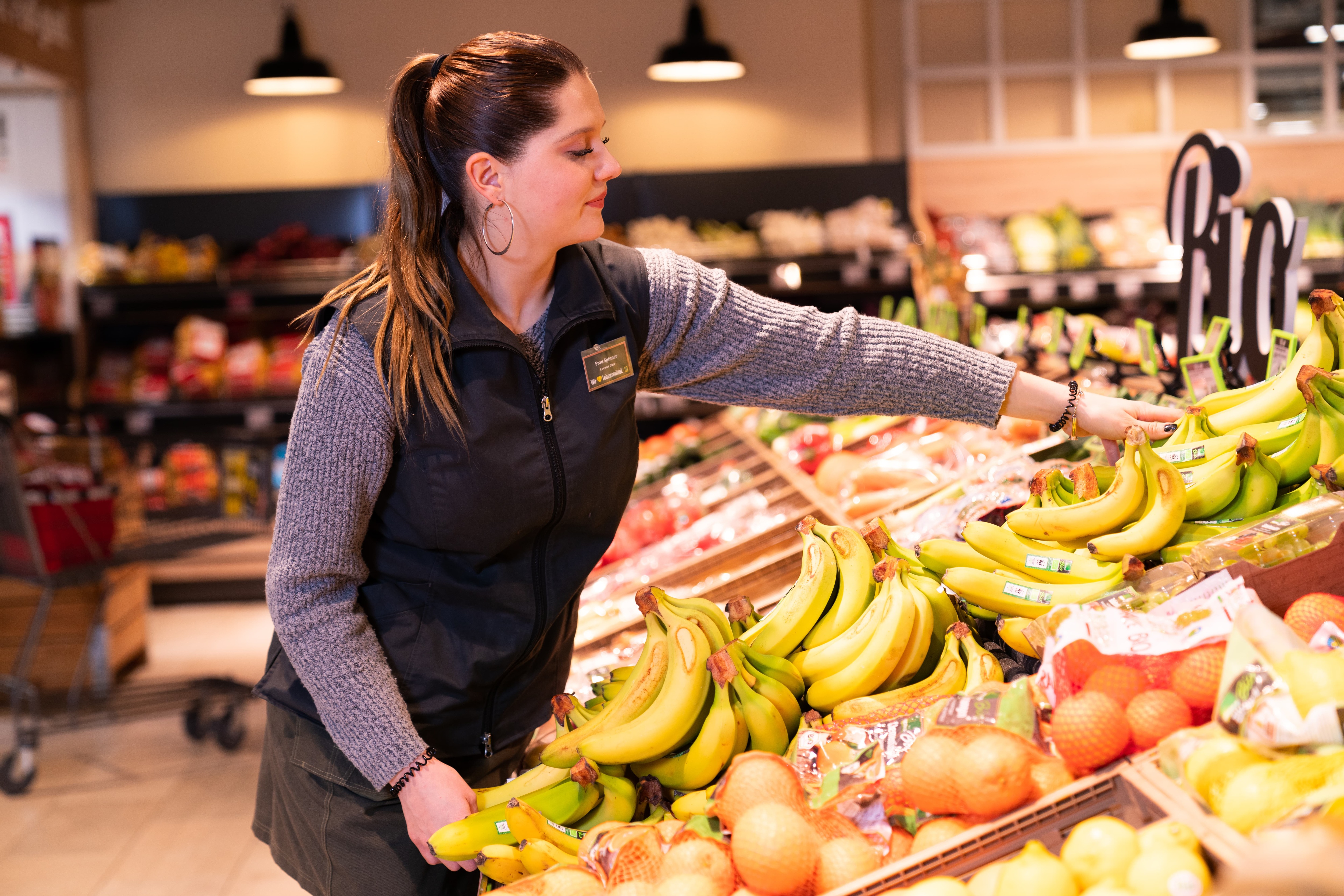 Eine Frau in grauem Wollpulli und schwarzer Weste greift ins Bananenregal. Sie trägt ein Namensschild von EDEKA.
