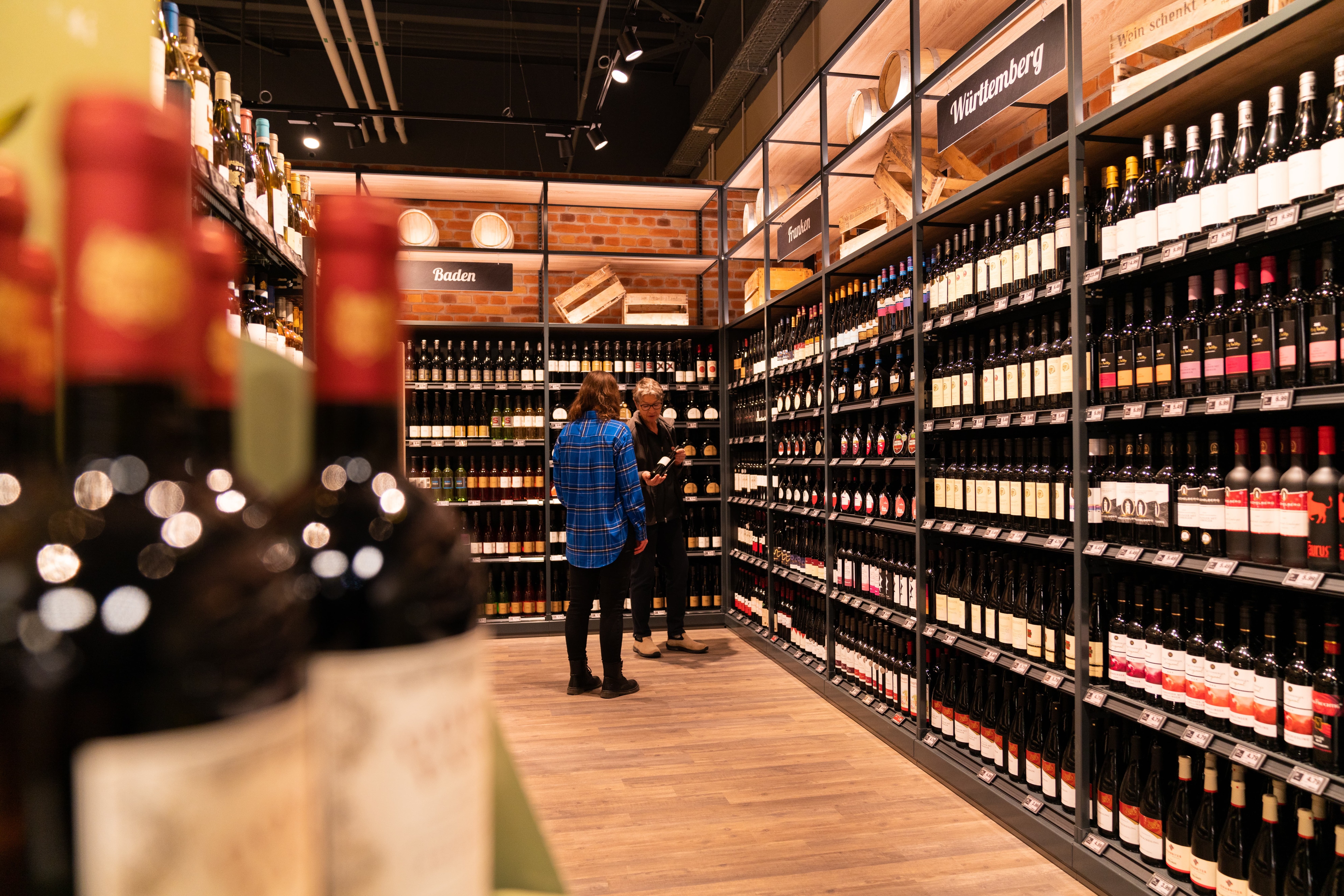 An einem Weinregal stehen zwei Frauen. Eine zeigt der anderen eine Flasche Rotwein.