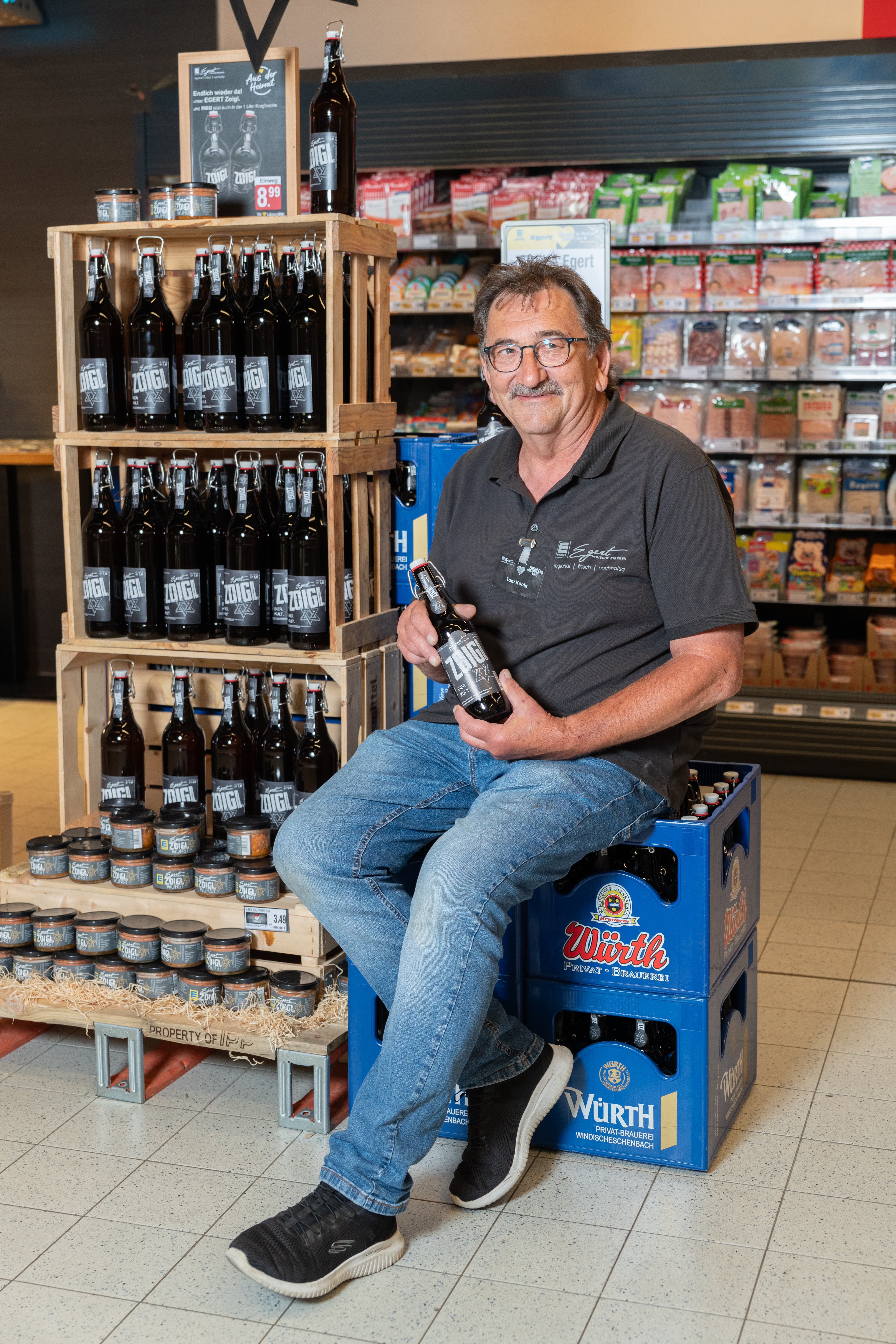 Ein lächelnder Mitarbeiter sitzt auf zwei Kisten Bier und hält eine Flasche Bier in den Händen.