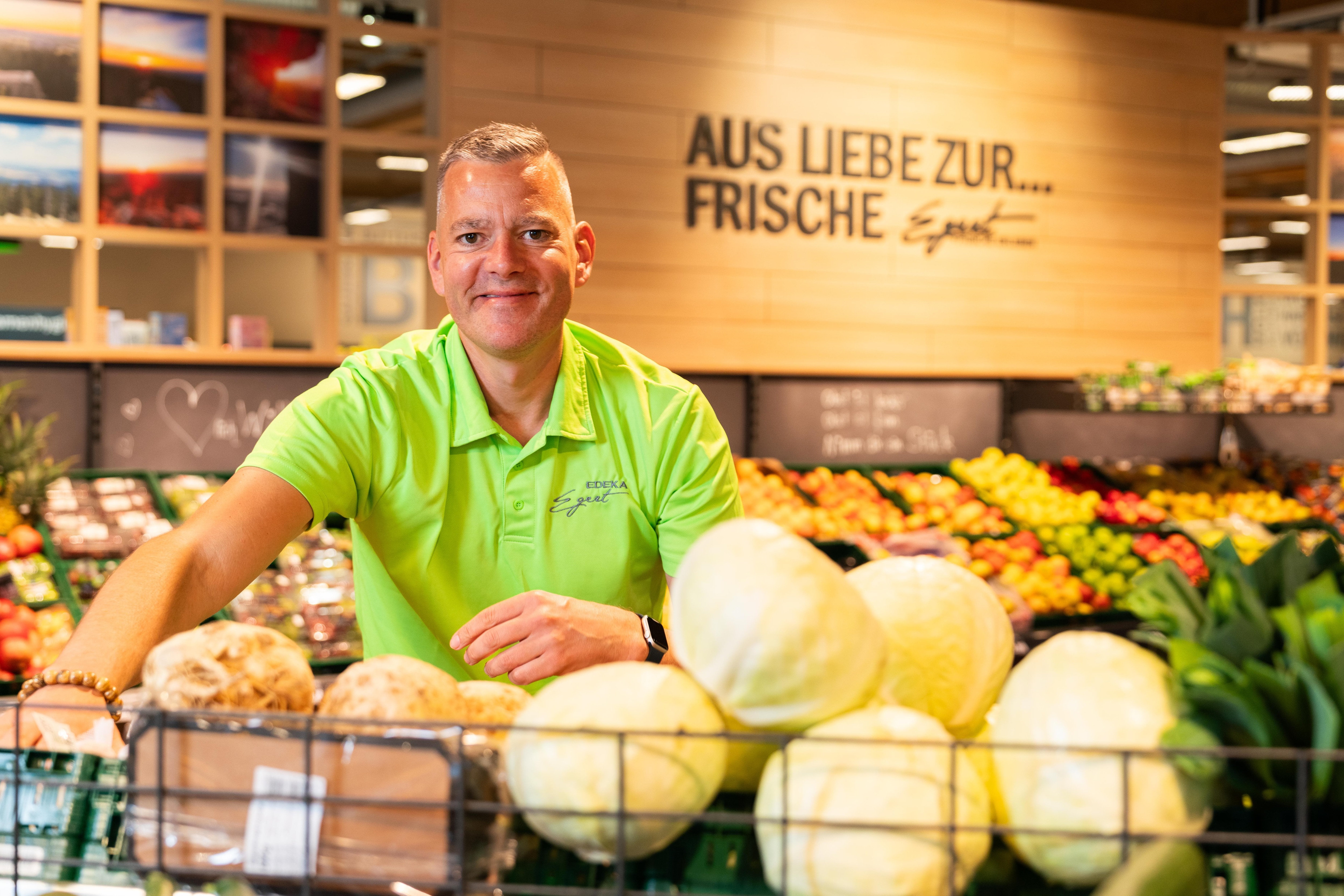 Der lächelnde Inhaber eines EDEKA-Marktes sortiert Produkte in der Obst- und Gemüseabteilung.