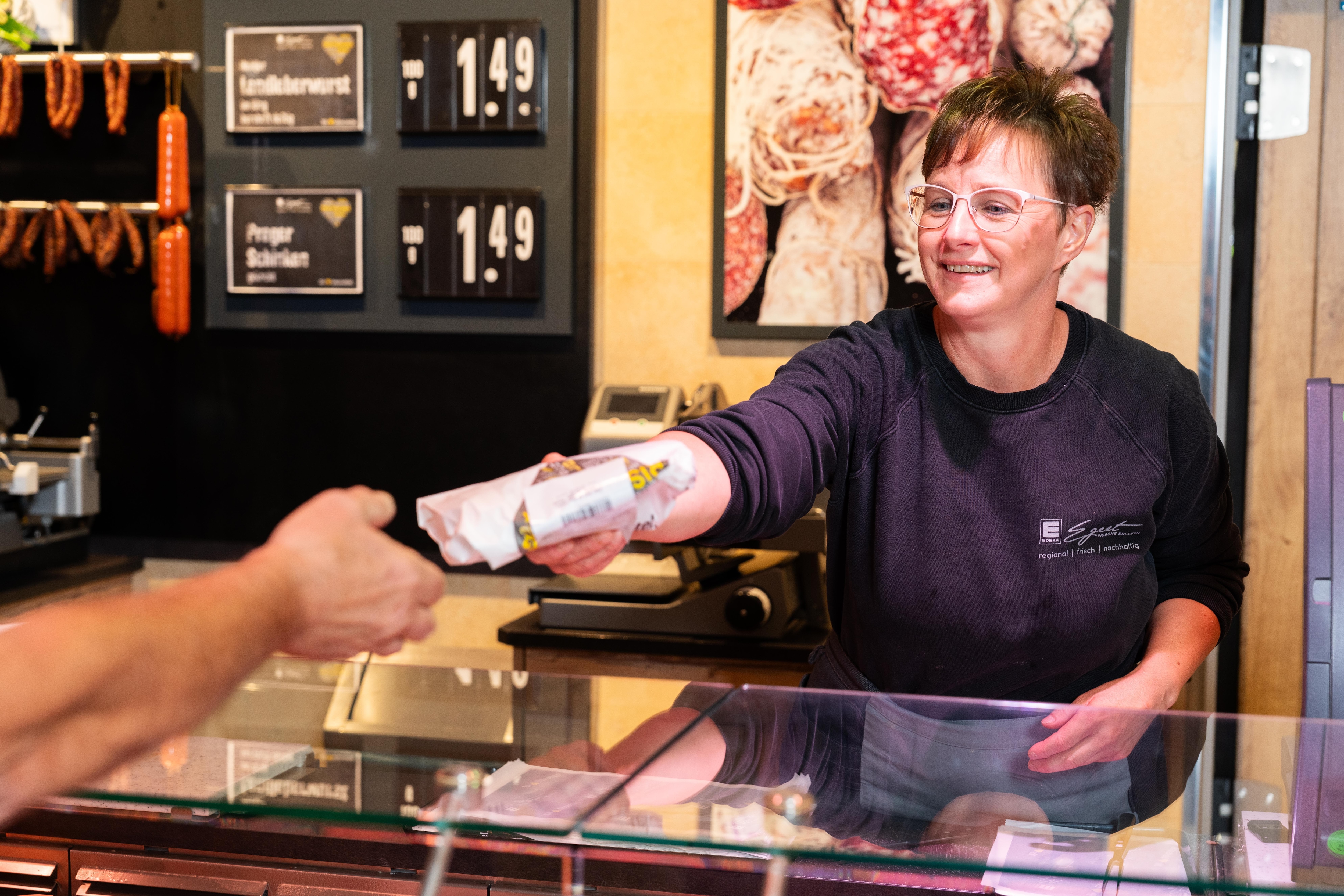 Eine lächelnde Mitarbeiterin der Bedientheke reicht einem Kunden ein Produkt über den Tresen.