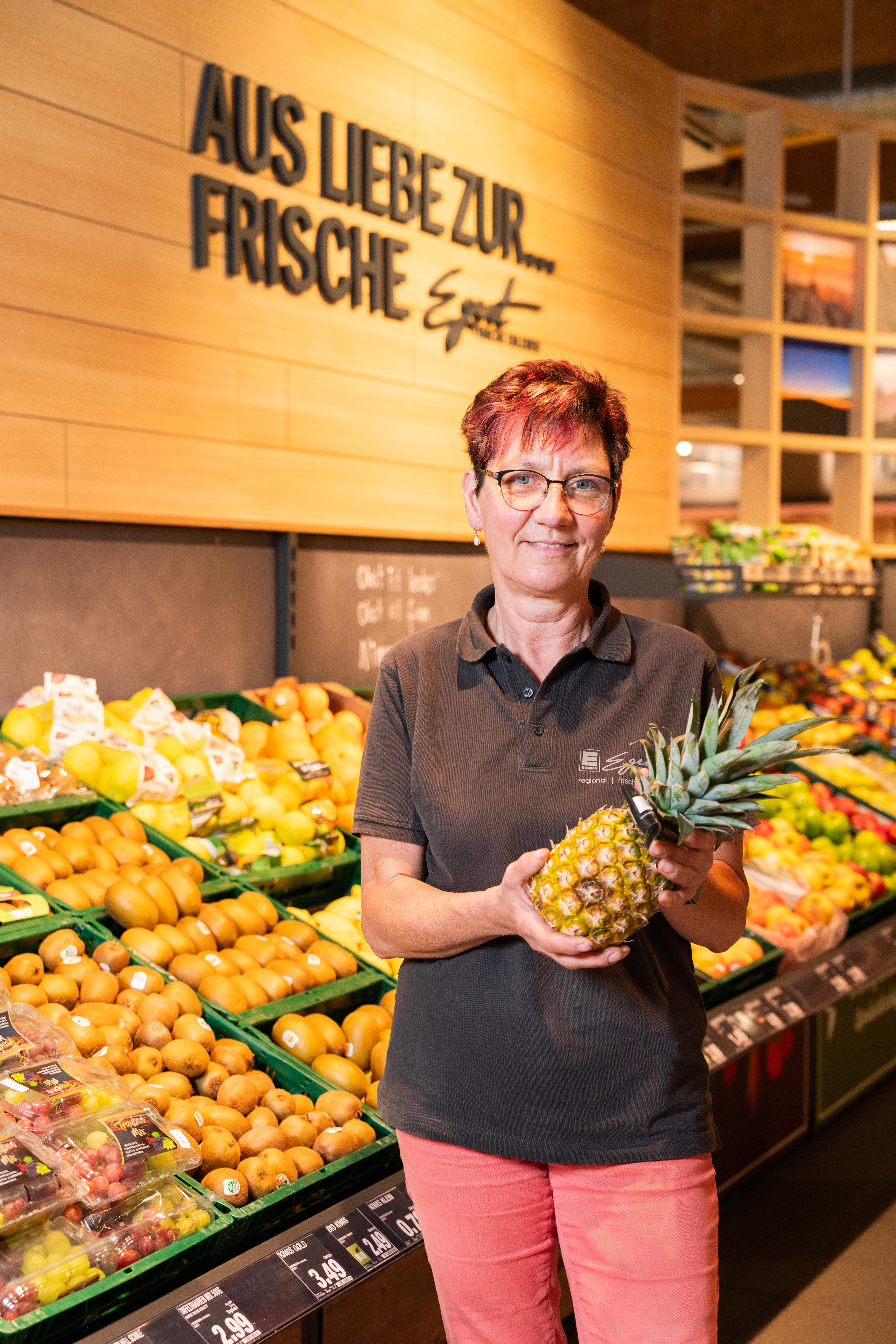 Eine lächelnde Mitarbeiterin steht in der Obstabteilung eines EDEKA-Marktes und hält eine Ananas in den Händen.