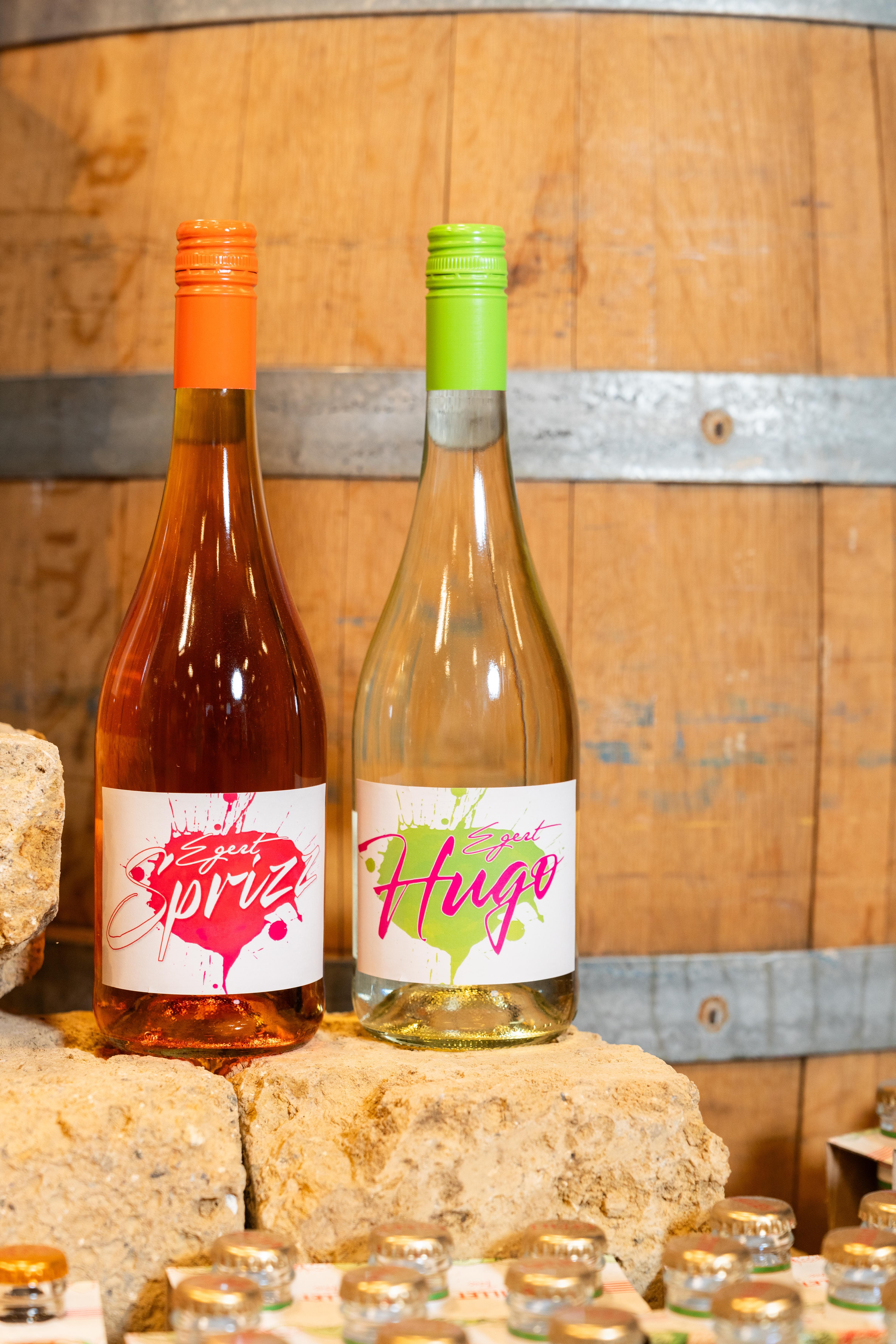 Eine Flasche Spritz und eine Flasche Hugo stehen auf einem Stein vor einem Eichenfass.