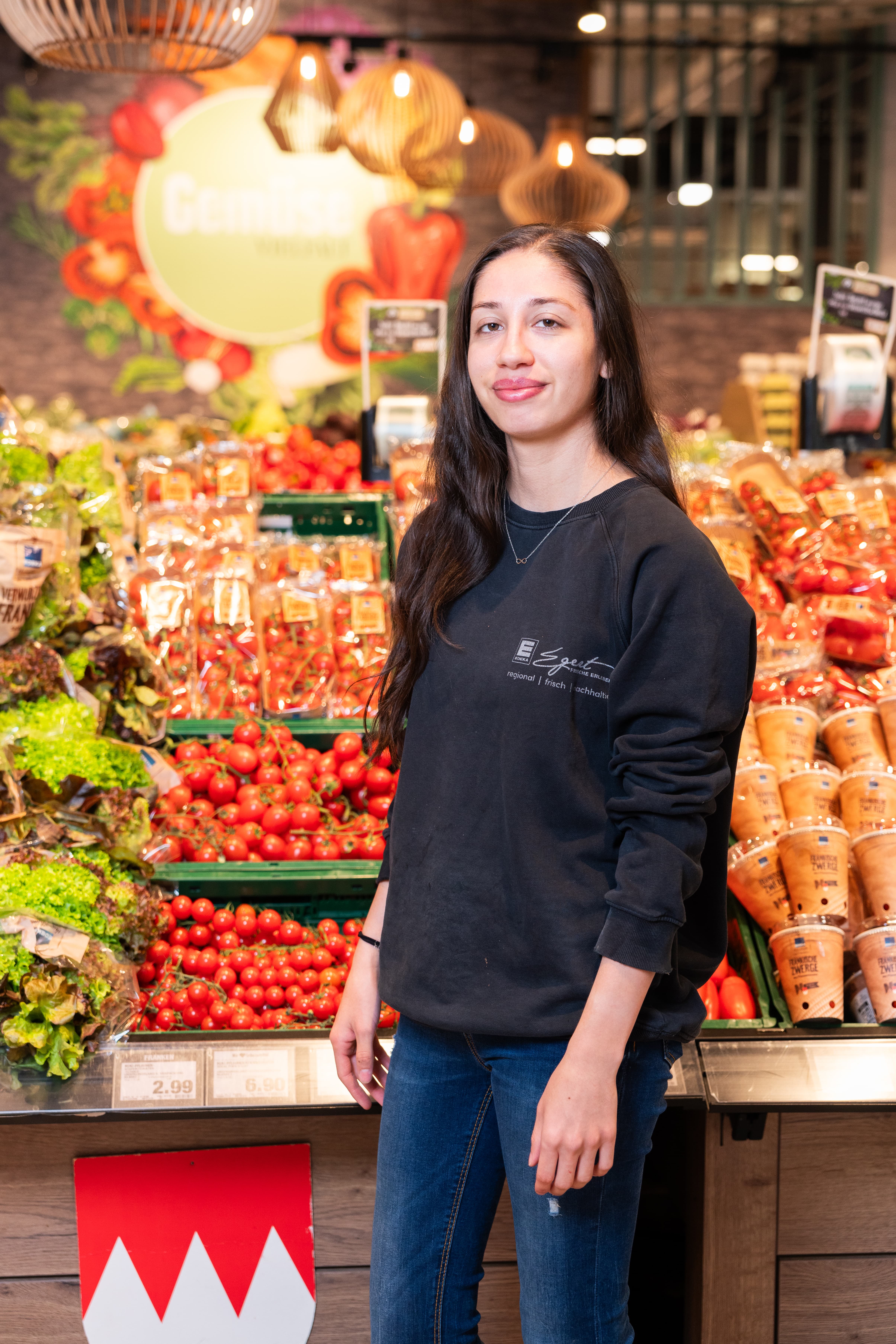 Eine lächelnde Mitarbeiterin eines EDEKA-Marktes steht in der Gemüseabteilung.
