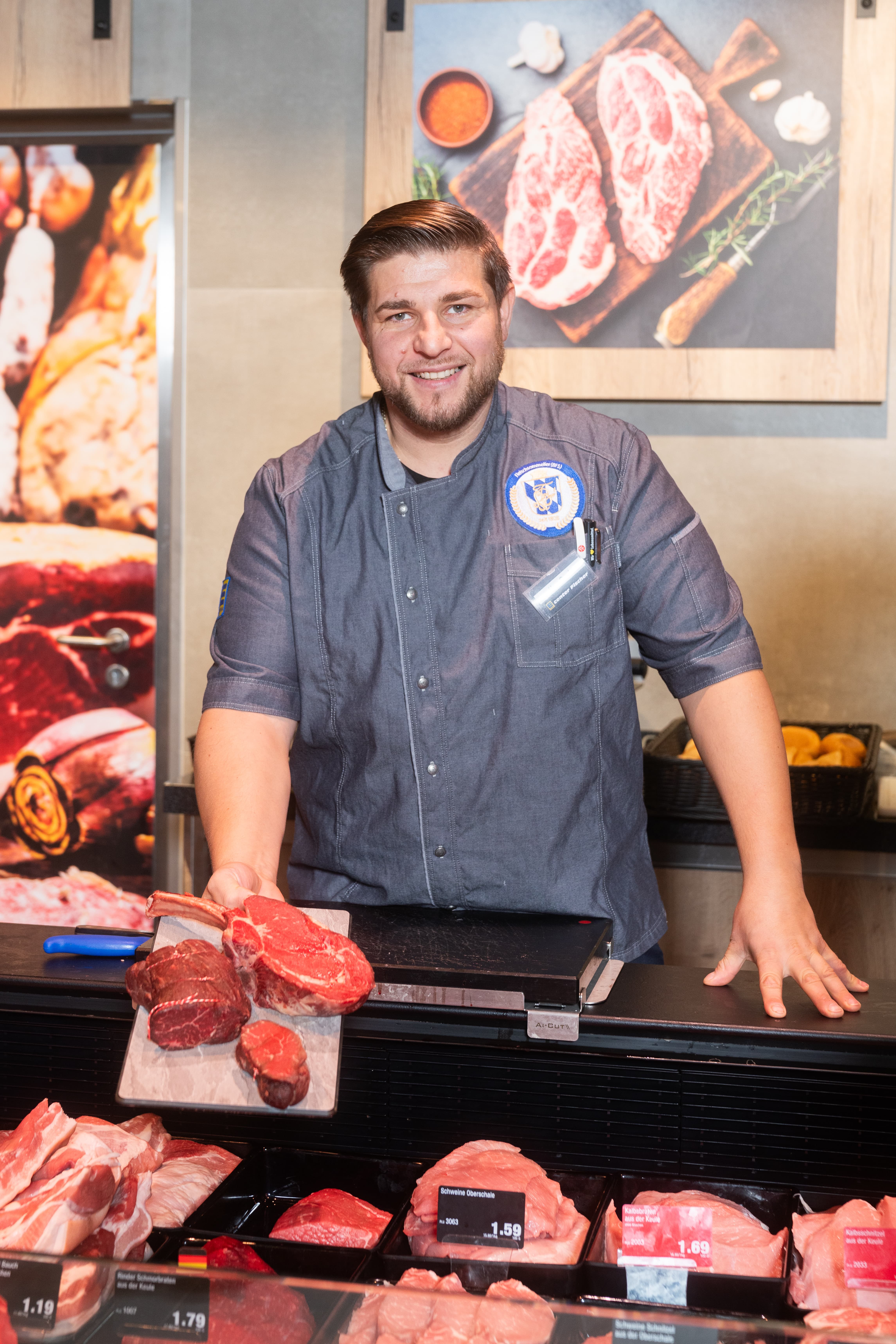 Ein Mann in blauem Fleischerkittel steht hinter einem Tresen hält ein Tablett mit drei Stücken Fleisch nach vorne