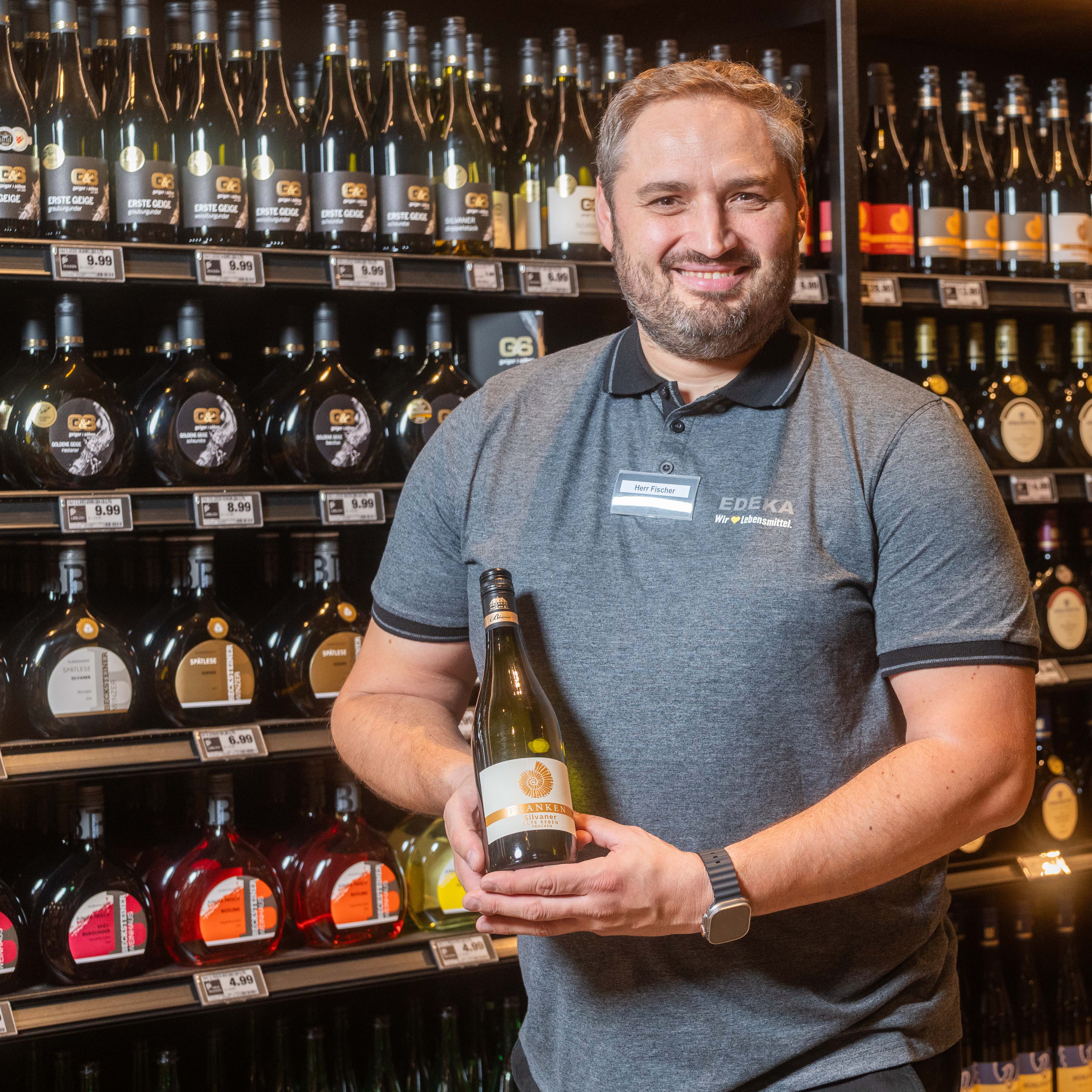 Herr Fischer steht vor dem Weinregal mit einer Flasche Wein in Händen. Er trägt ein graues Poloshirt mit EDEKA-Logo.