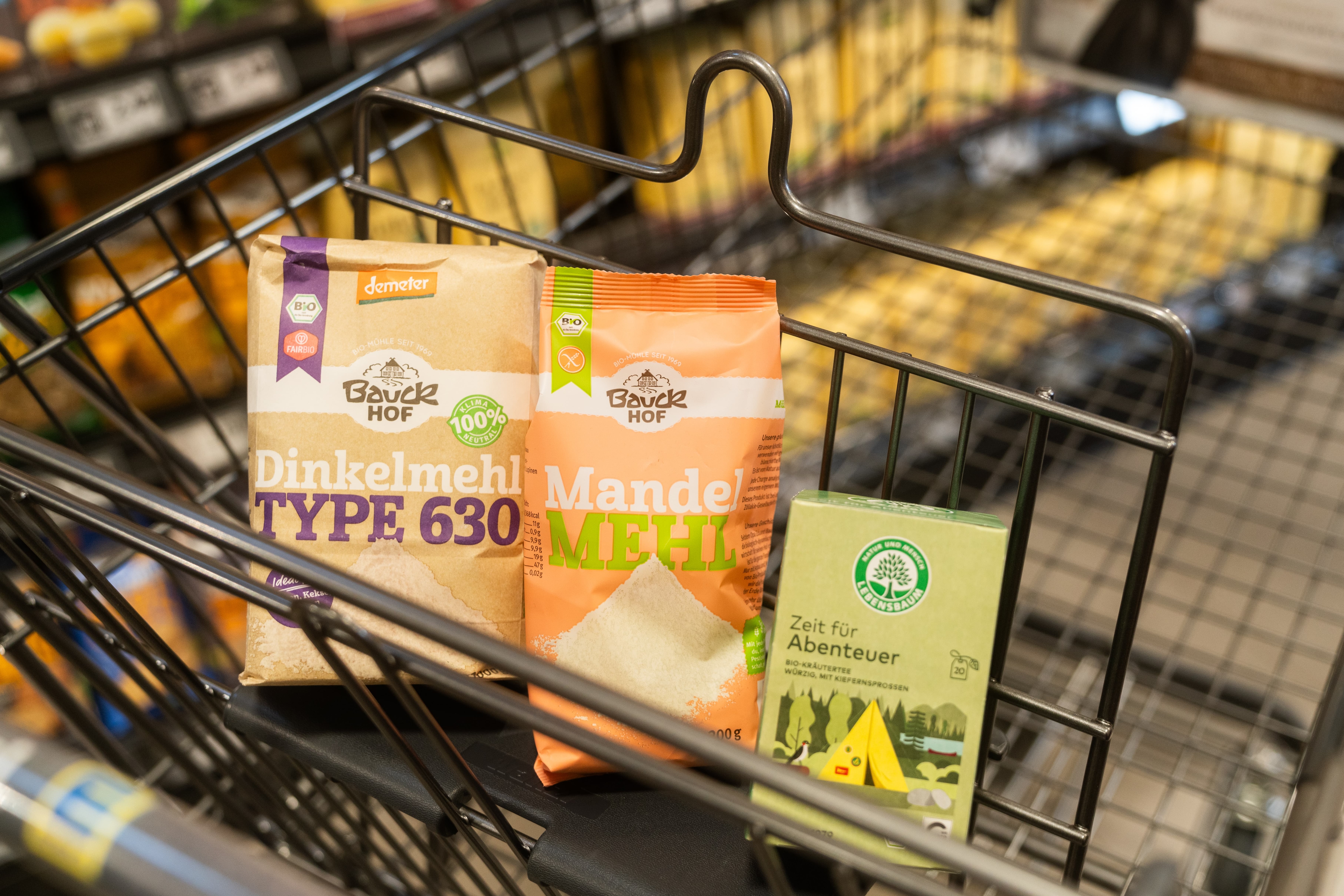 In einem Einkaufswagen liegen Dinkelmehl, Mandelmehl und eine Packung Bio-Krätertee.