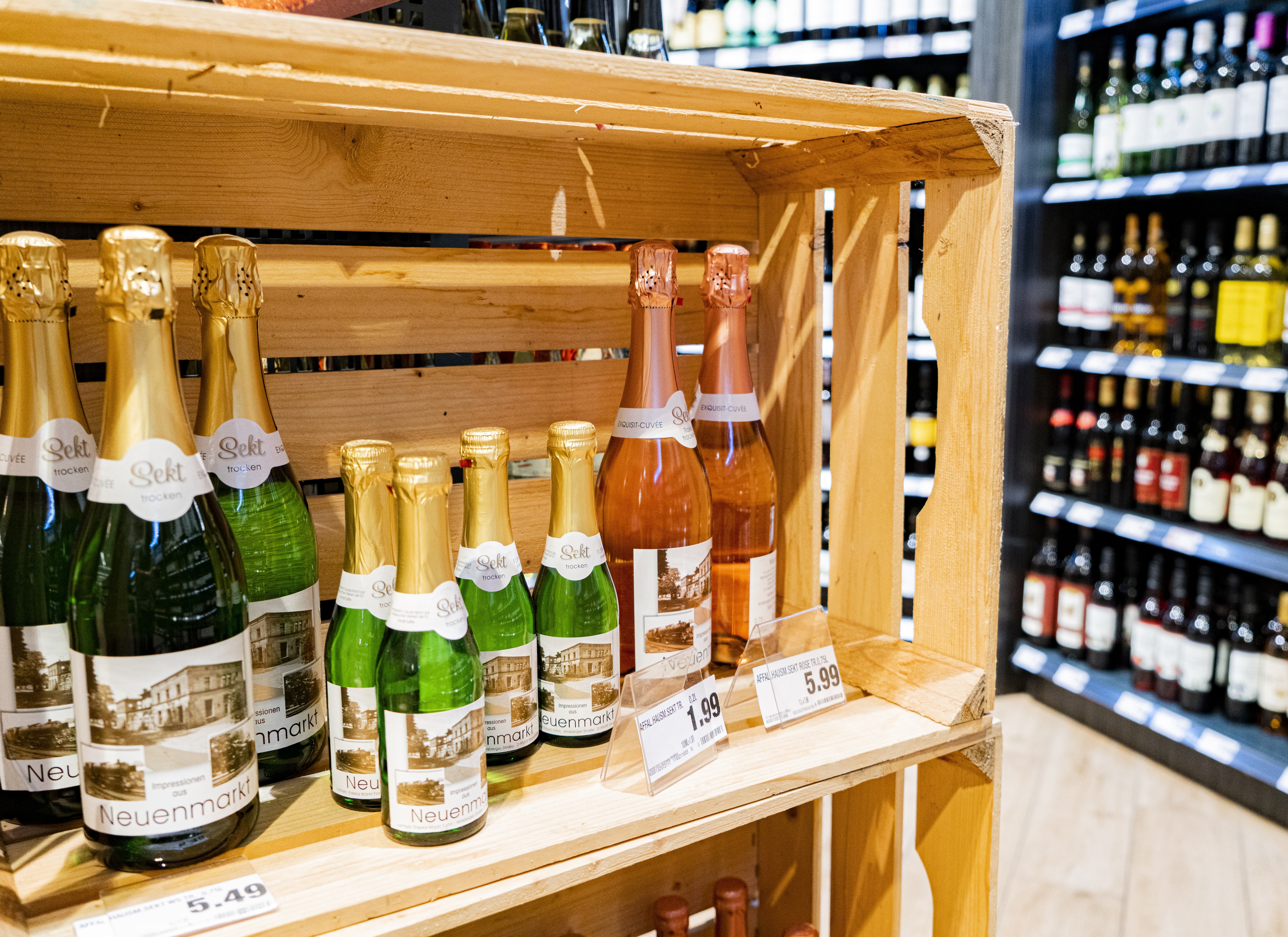 Mehrere Sektflaschen in einer Holzkiste, im Hintergrund die Weinabteilung eines EDEKA-Markts.