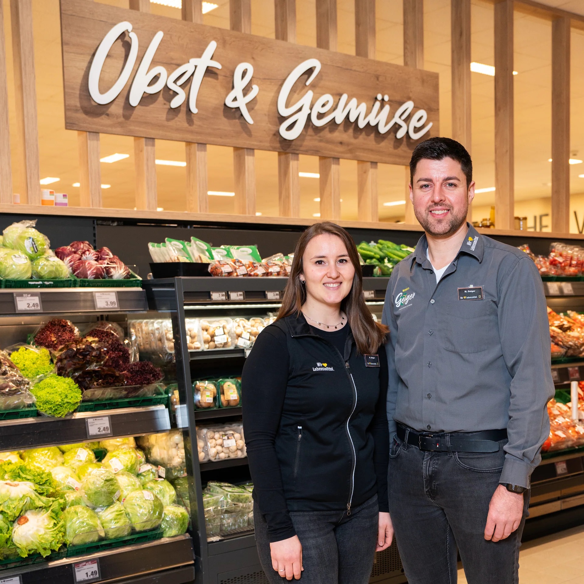 Eine Frau und Mann in Arbeitskleidung von Edeka stehen in einer Obst- & Gemüseabteilung und lächeln.