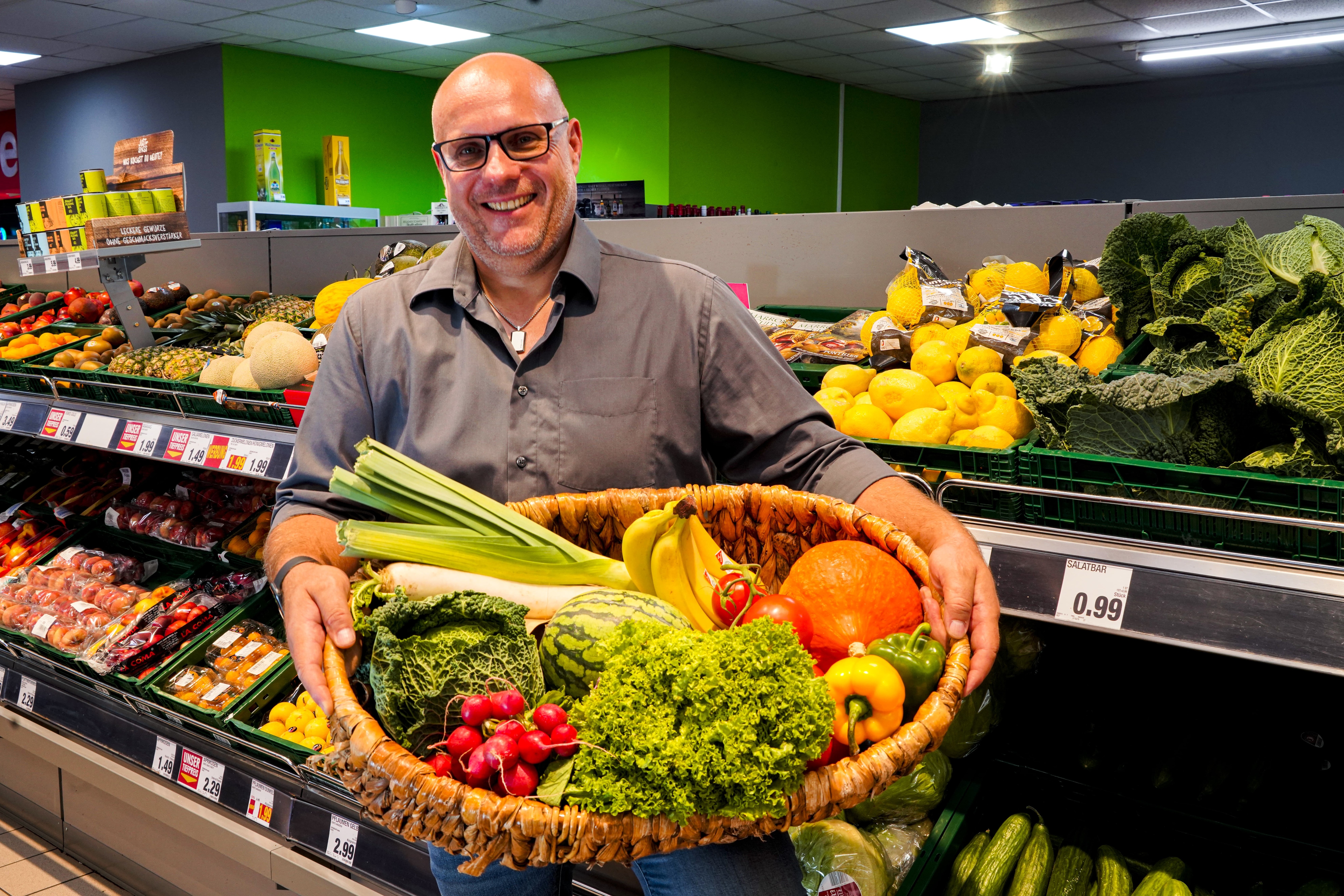 Inhaber des Edeka Marktes steht in der Gemüße- und Obstabteilung. Er hat einen Korb mit frischen Obst- und Gemüse in den Händen und lächelt.