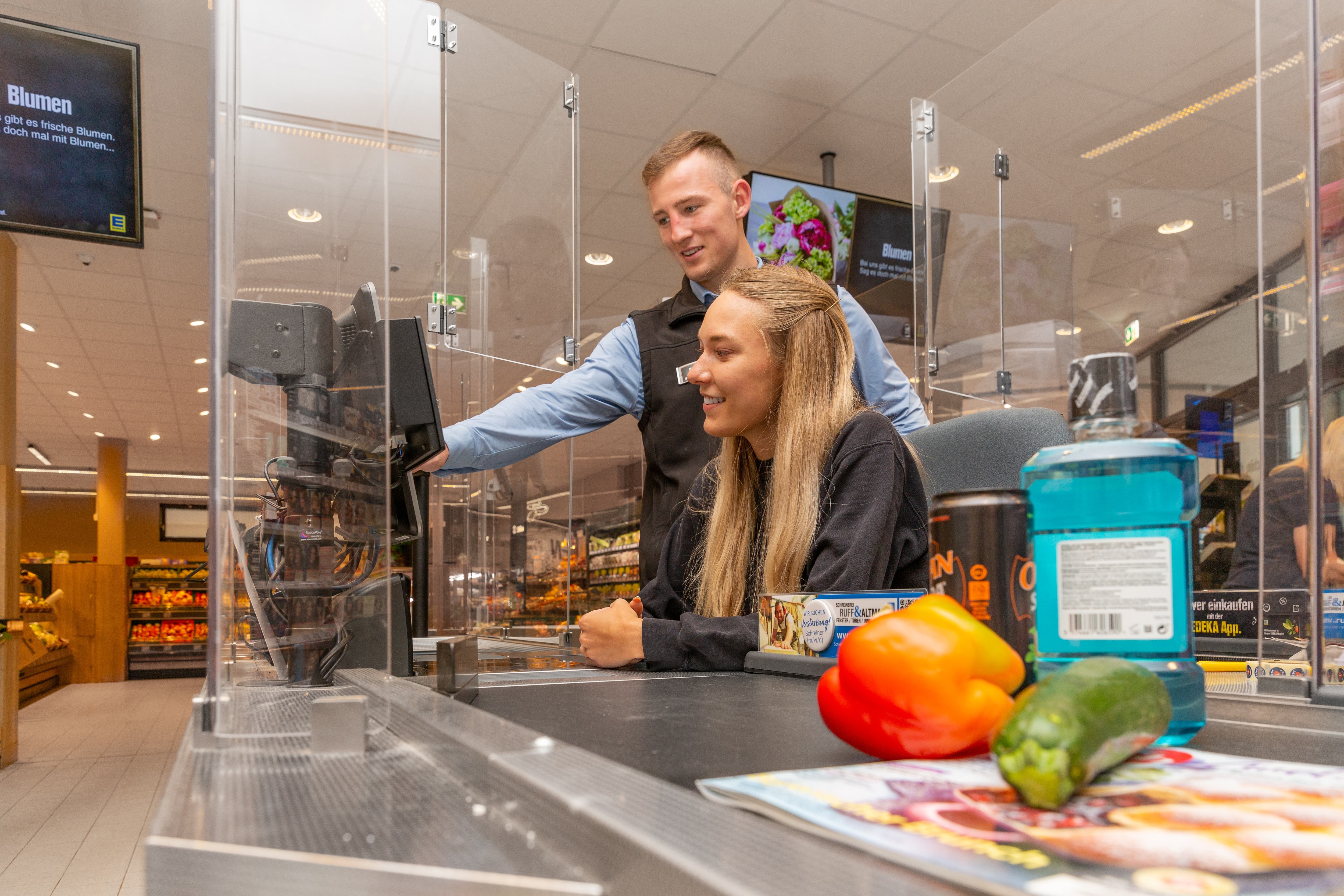 Ein Mitarbeiter eines Edeka-Marktes erklärt einer Auszubildenden etwas an der Kasse.