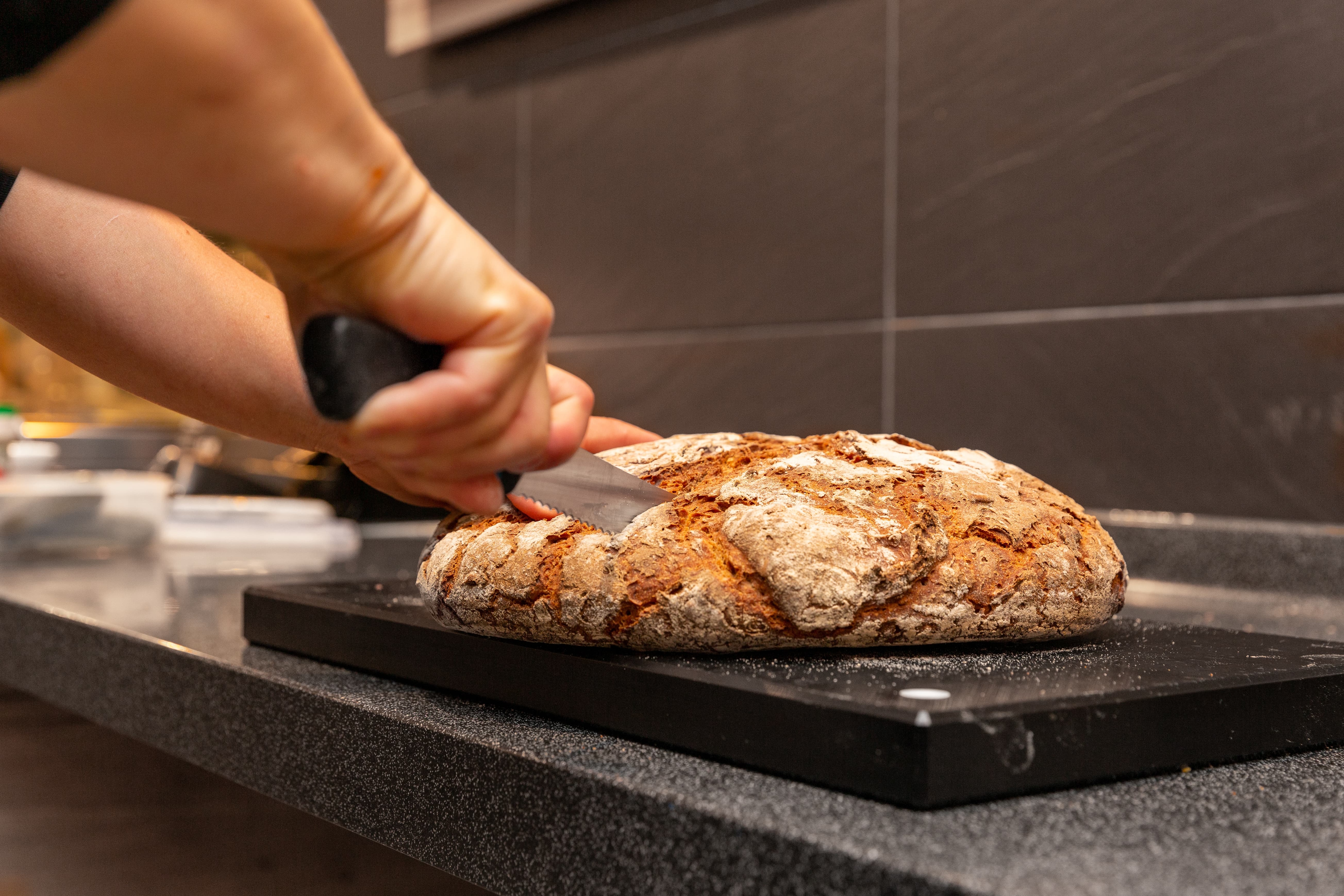 Ein Mitarbeiter schneidet ein Leibbrot mit einem Sägemesser auf einem schwarzen Schneidebrett.