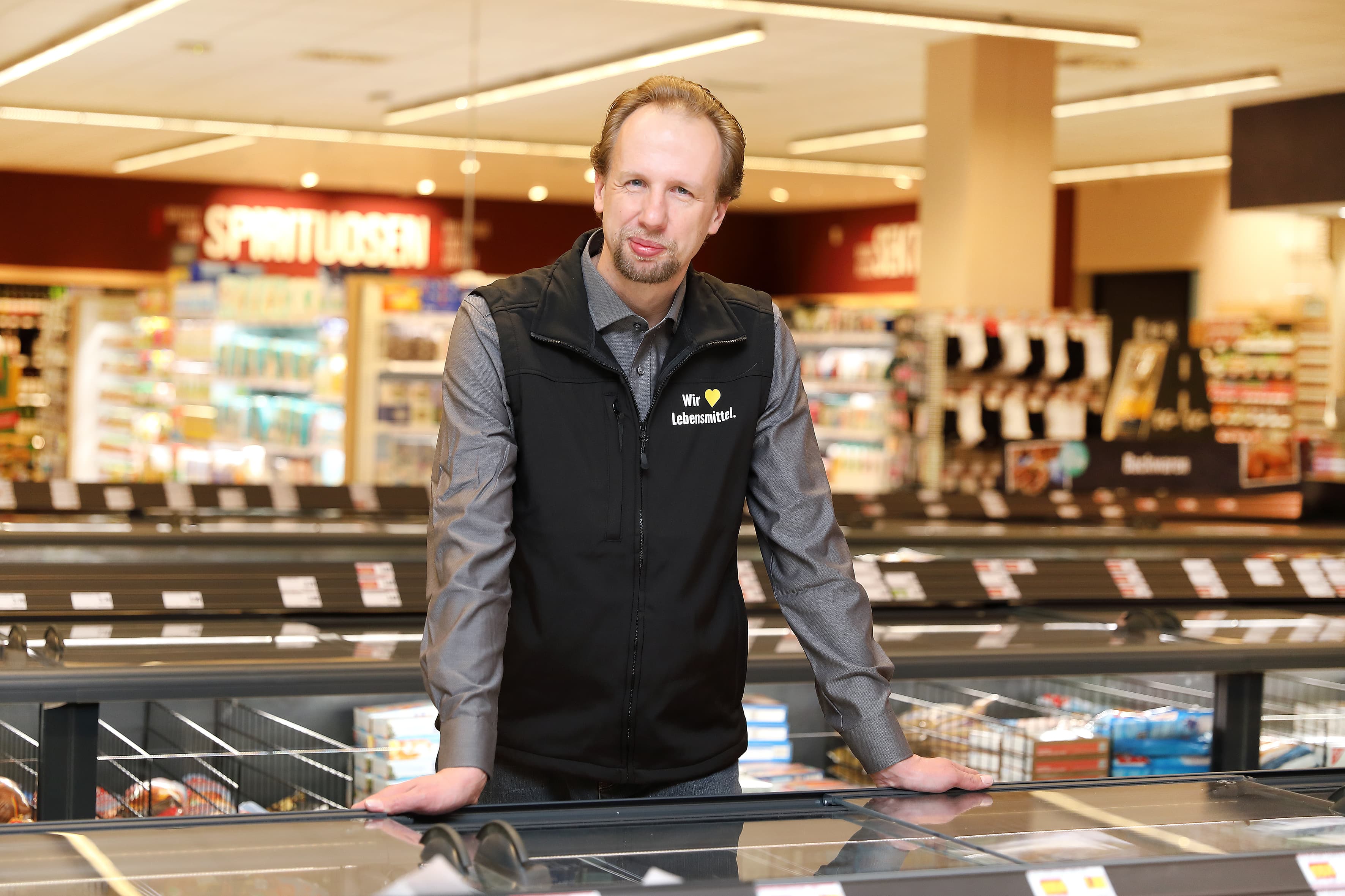 Der Marktleiter eines Edeka-Marktes lehnt sich auf eine Tiefkühltruhe und lächelt in die Kamera.