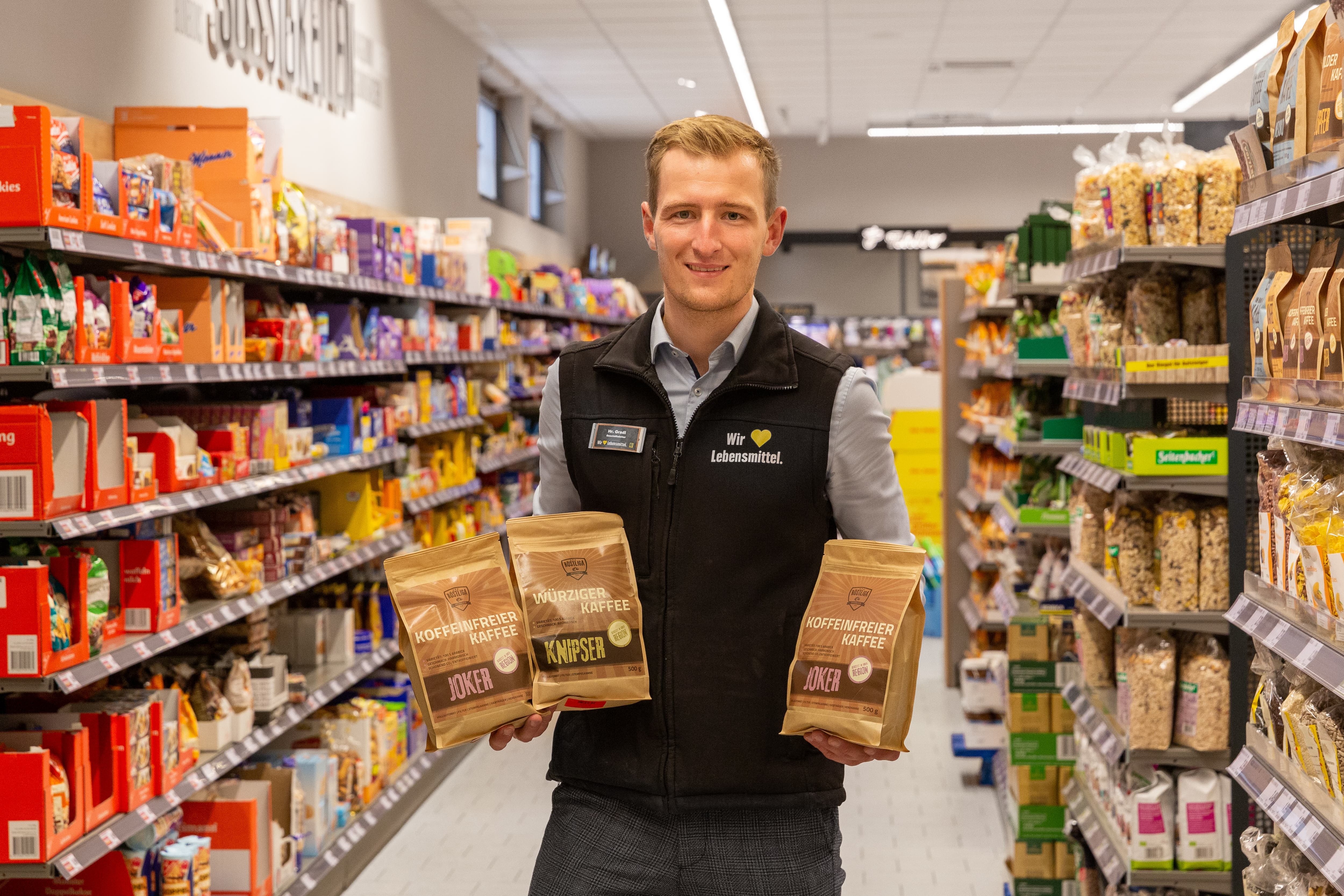 Ein lächelnder Mitarbeiter steht im Gang eines Edeka-Marktes und hält drei verschiedene Packungen regionalen Kaffees in den Händen.