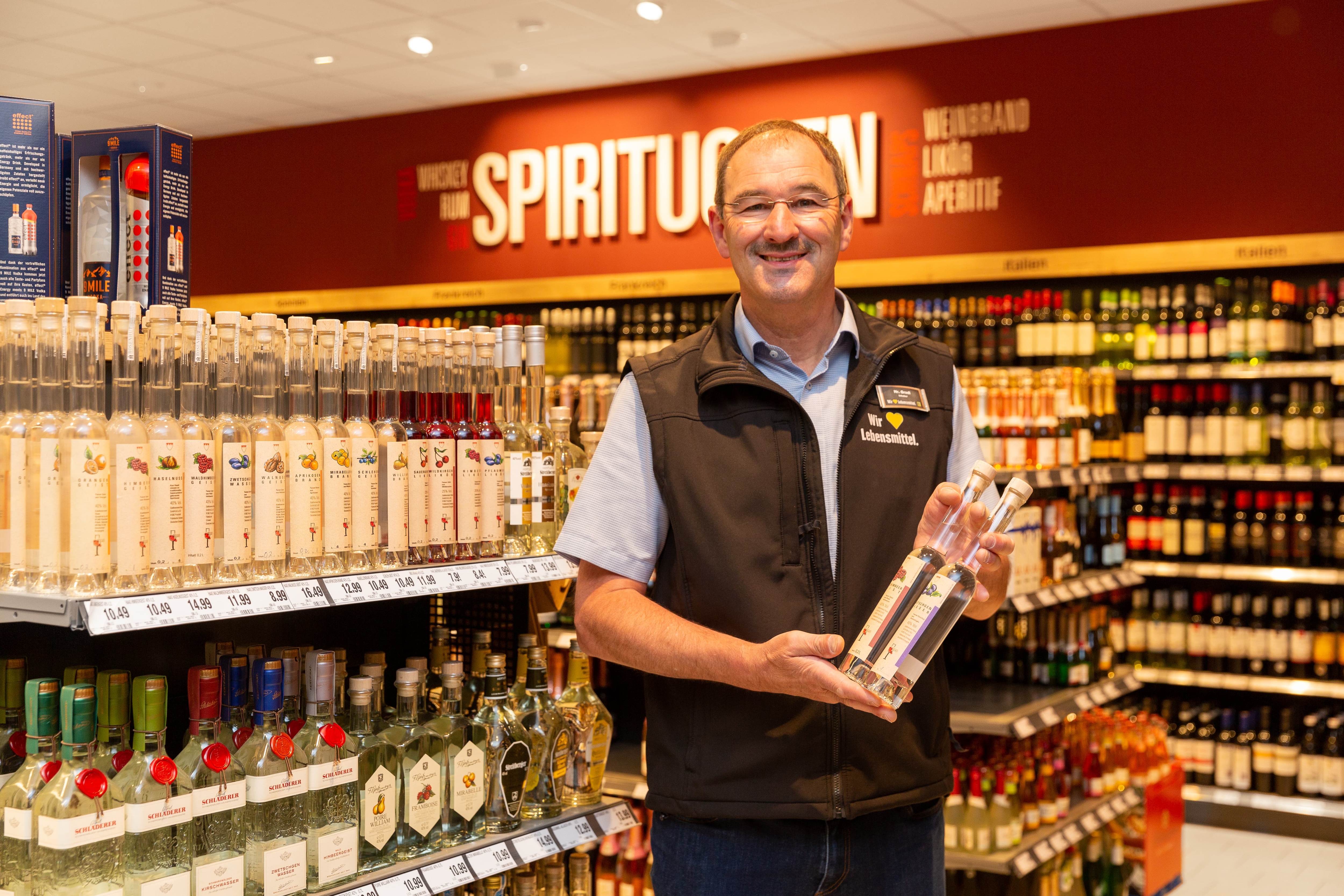 Ein lächelnder Mitarbeiter eines Edeka-Marktes steht in der Spirituosenabteilung präsentiert zwei Flaschen Weinbrand.
