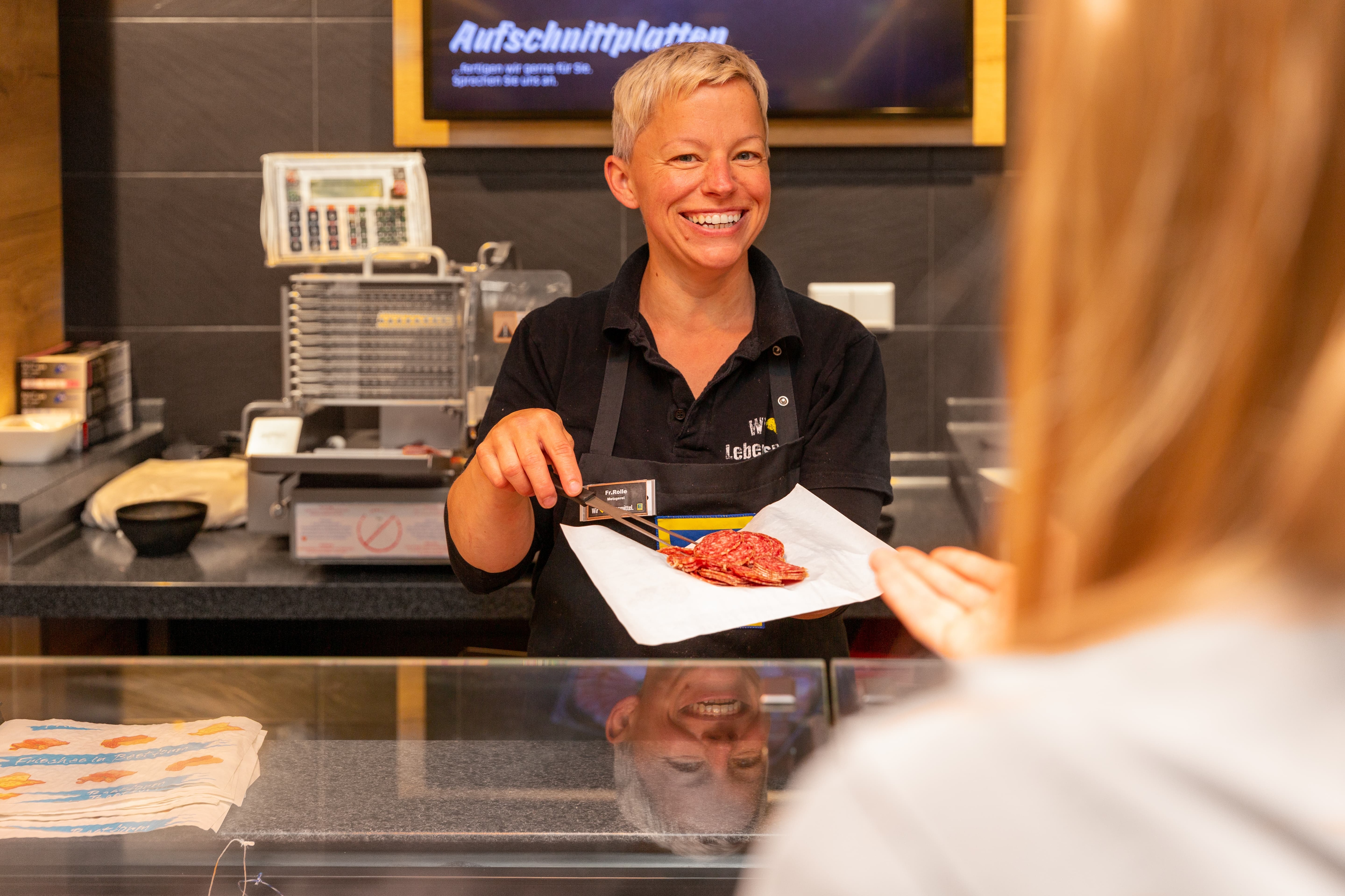 Eine lächelnde Mitarbeiterin der Fleischtheke eines Edeka-Marktes legt Wurstscheiben auf ein Stück Papier.