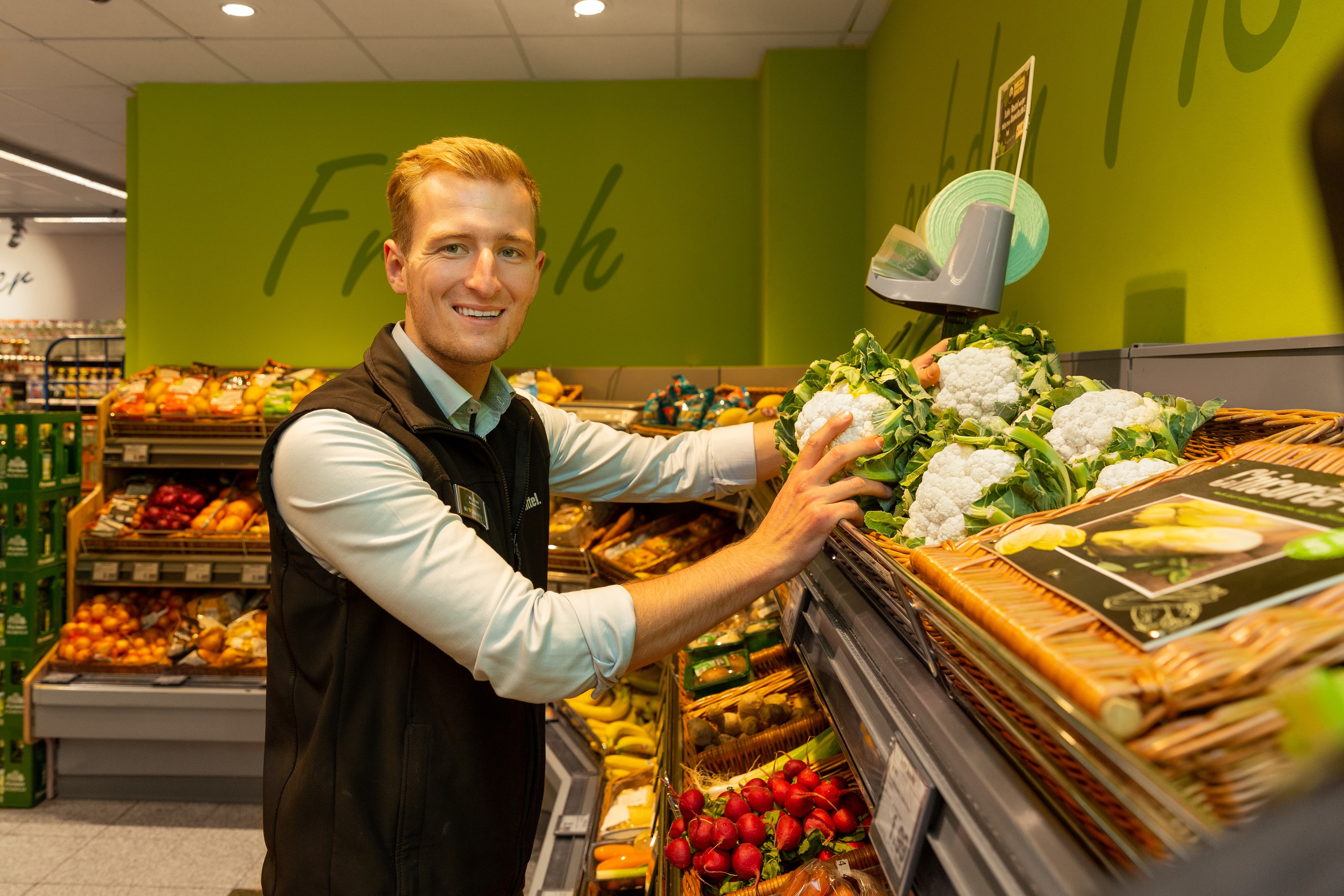 Ein lächelnder Mitarbeiter eines Edeka-Marktes verräumt Blumenkohl in ein Regal.
