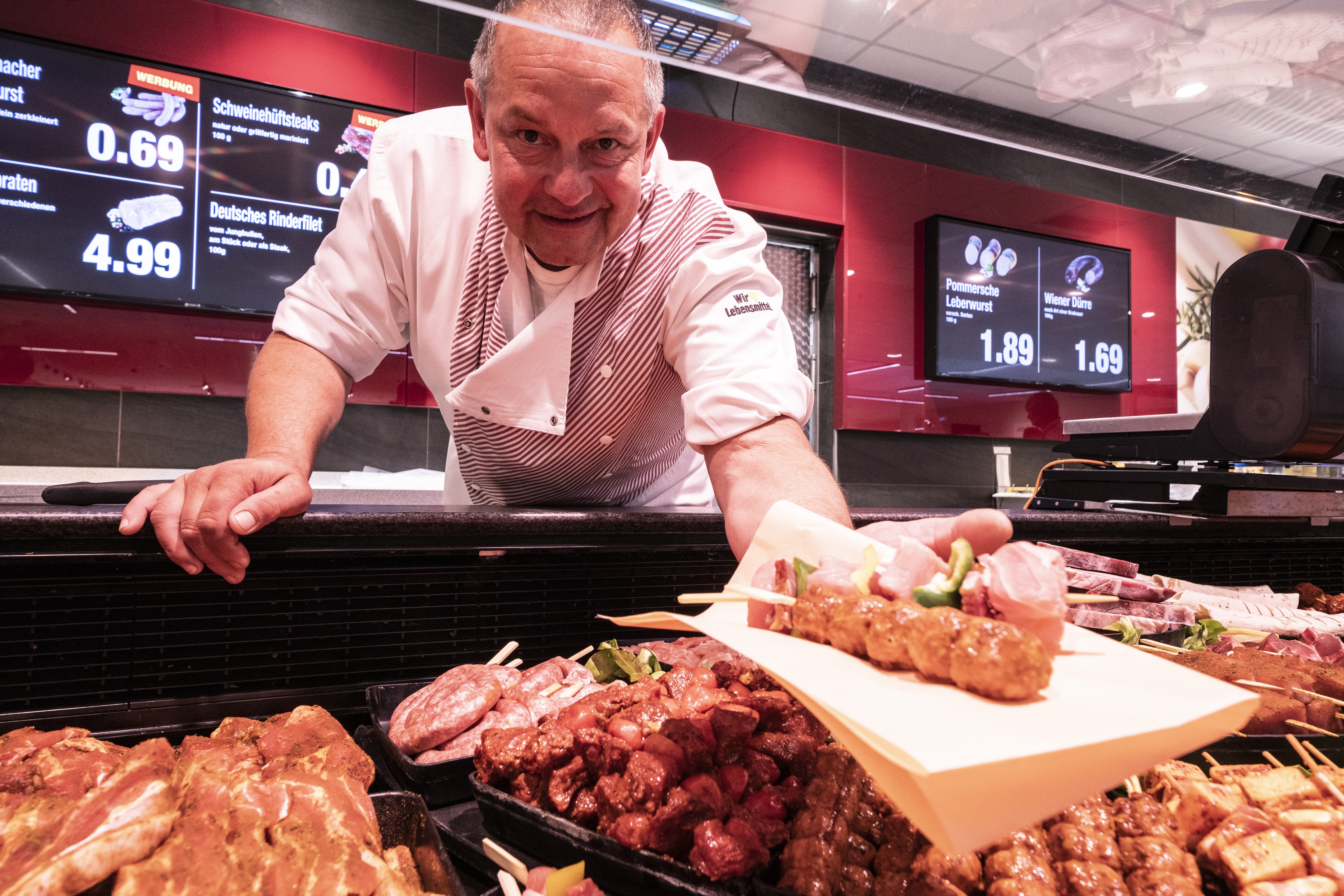 An der Bedientheke eines EDEKA-Markts hält ein Mitarbeiter lächelnd zwei Fleischspieße auf einer Serviette in die Kamera.