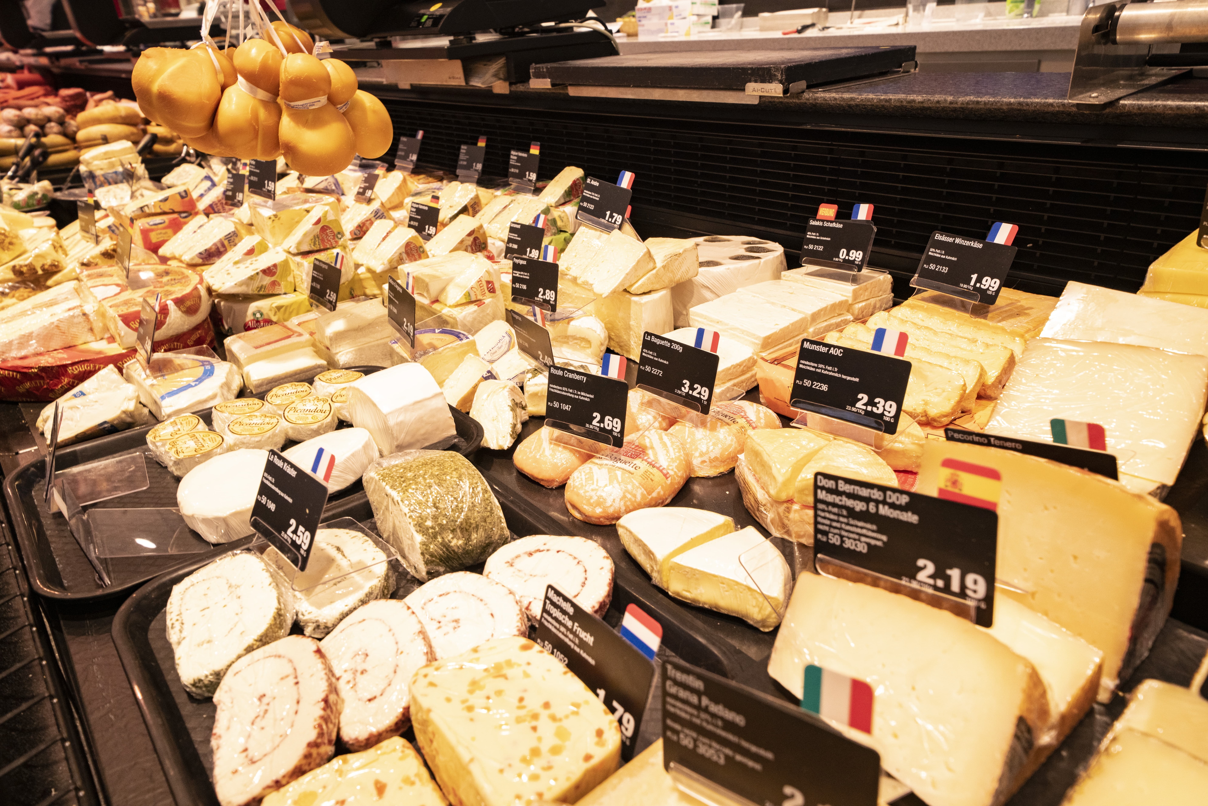 Käseauslage einer Bedienungstheke mit vielen verschiedenen Käsesorten und Preisschildern. Geräucherter Mozzarella hängt an Kordeln.