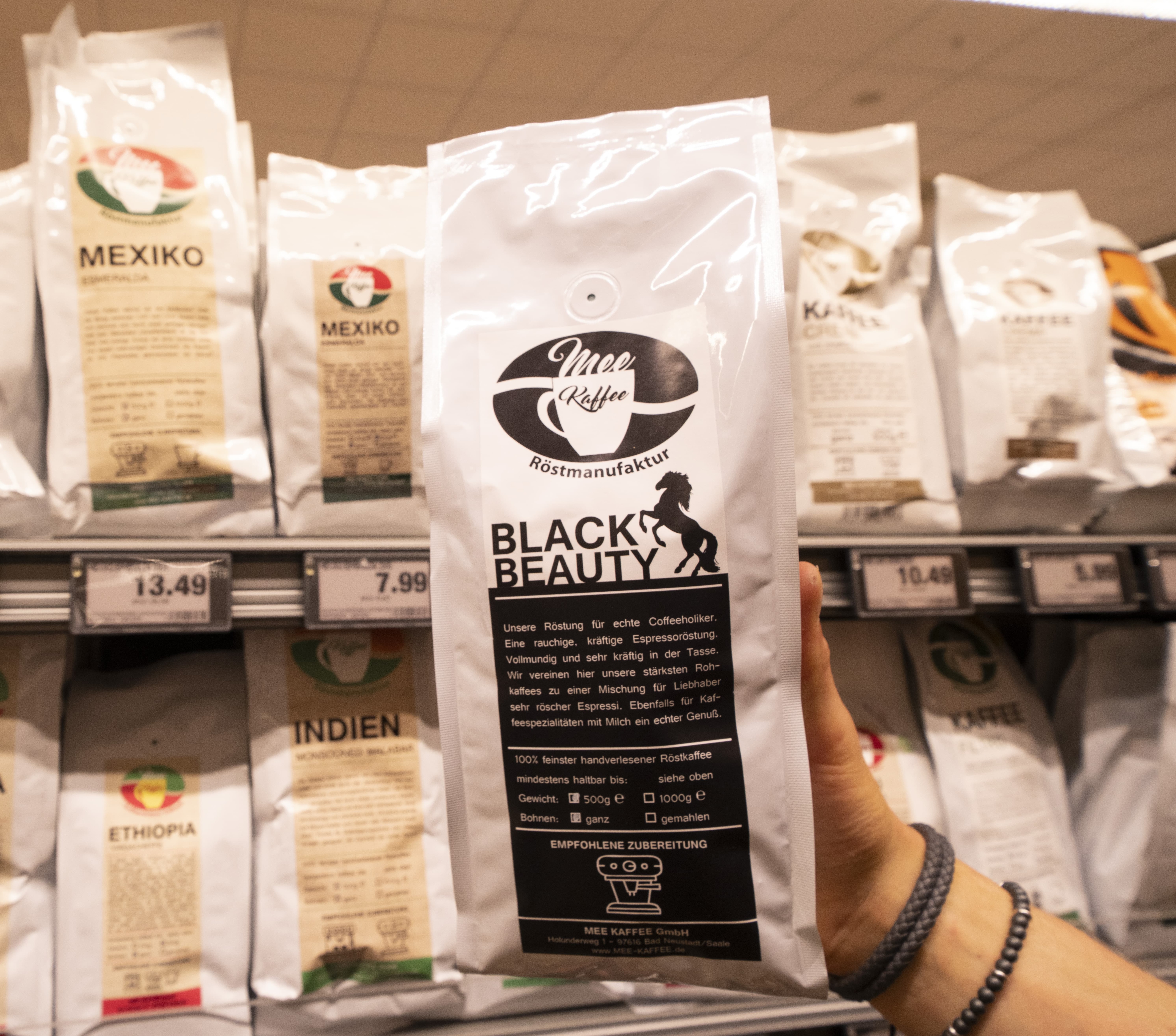 Eine Hand hält eine Packung Kaffee mit der Aufschrift „Black Beauty“ hoch, im Hintergrund ein Regal mit weiteren Packungen und verschiedenen Etiketten.
