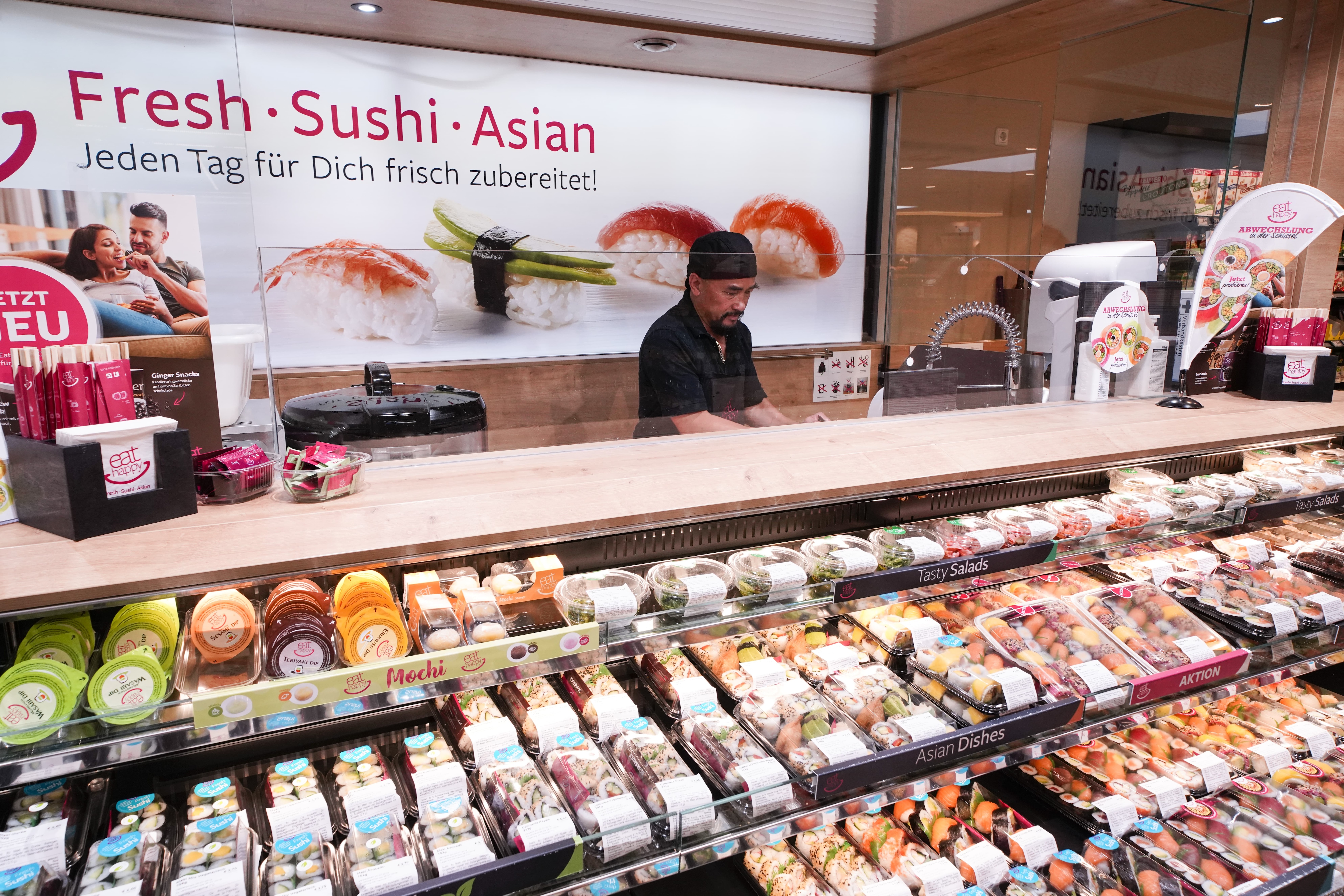 Die Sushiabteilung des EDEKA-Markts mit dem Banner: „Fresh Sushi Asian. Jeden Tag für dich frisch zubereitet!“ und einem Mitarbeiter hinter der Theke.