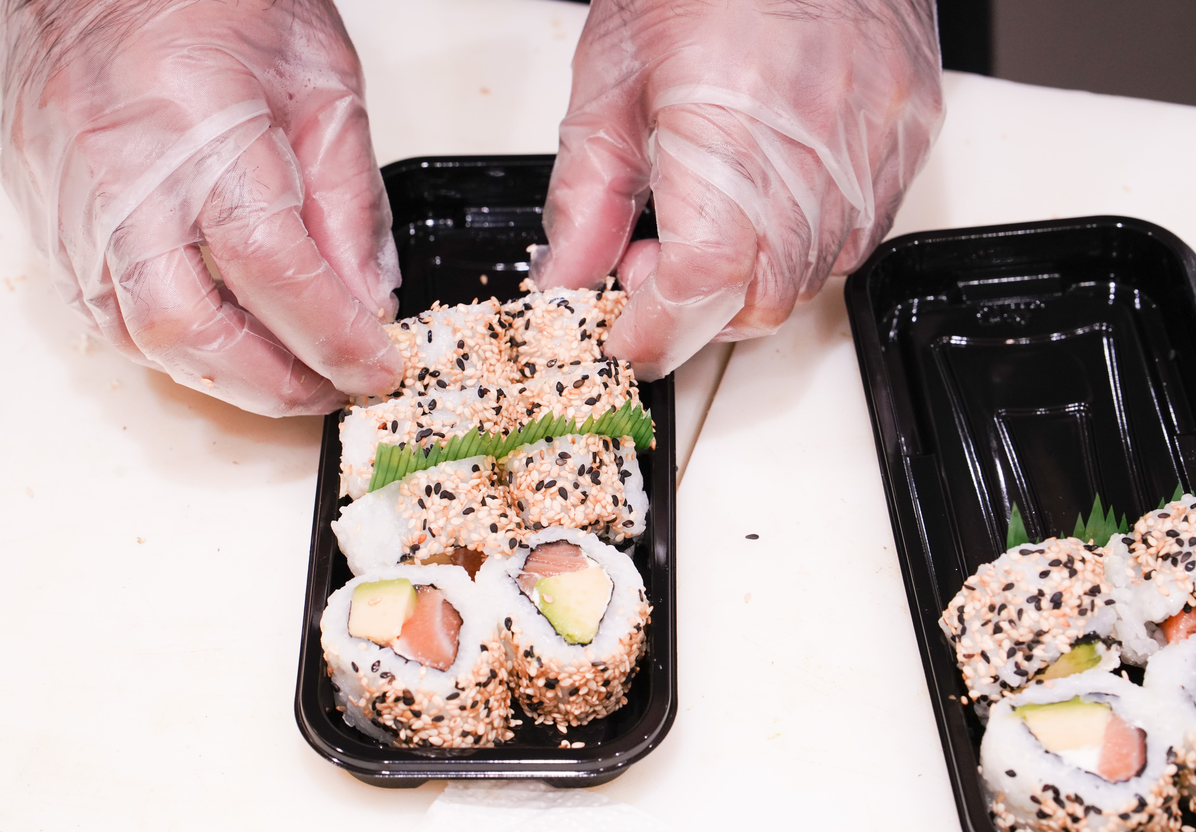 Zwei Hände mit Handschuhen ordnen Sushi in einer schwarzen Plastikschale an.