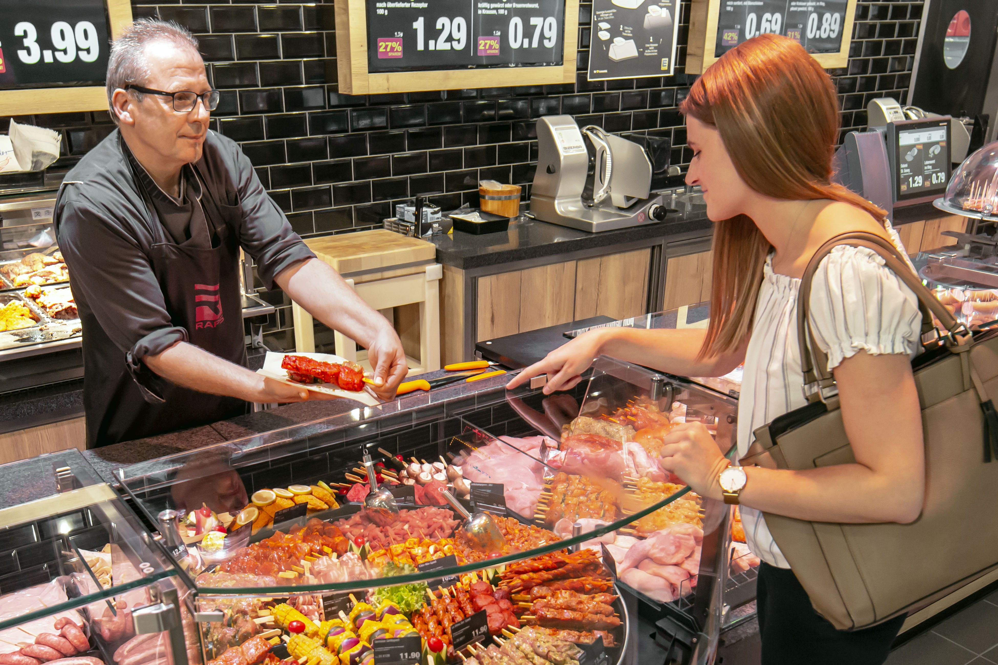 An der Bedientheke eines EDEKA-Markts zeigt ein Mitarbeiter einer Kundin lächelnd ein Stück Fleisch.