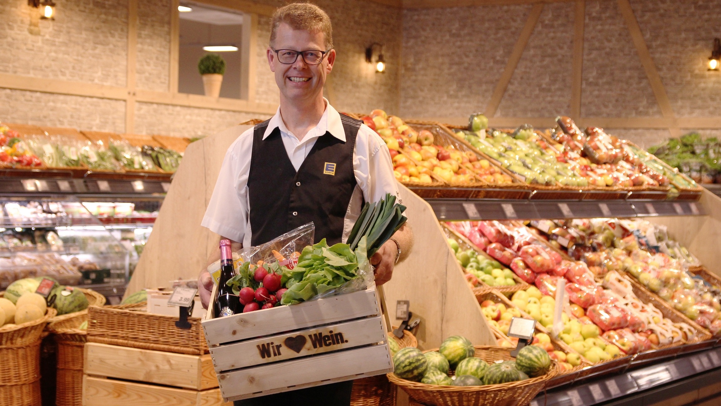 Der Markinhaber Hr. Körner steht lächelnd mit einer Gemüsekiste in der Hand in der Gemüse- und Obstabteilung.