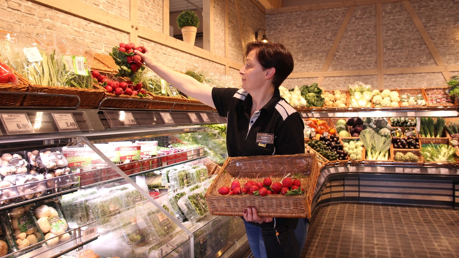 Die Marktleiterin Fr. Reimer räumt Radieschen ins Gemüseregal ein und steht seitlich zur Kamera.