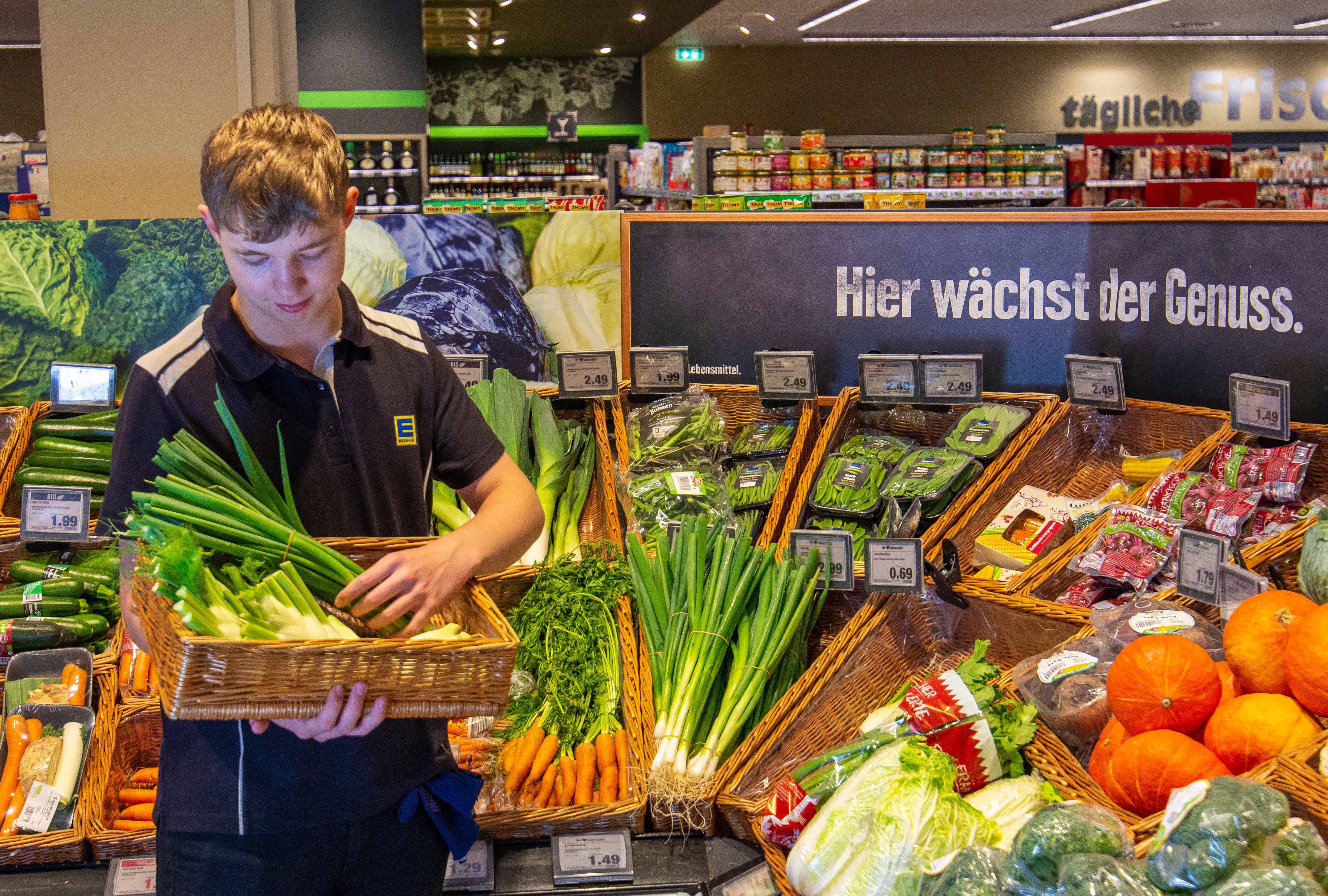 Ein Auszubildender hält einen Korb mit Lauchzwibeldn und Fenchel in der Hand. Er steht in der Gemüseabteilung und blickt auf den Korb hinhab.