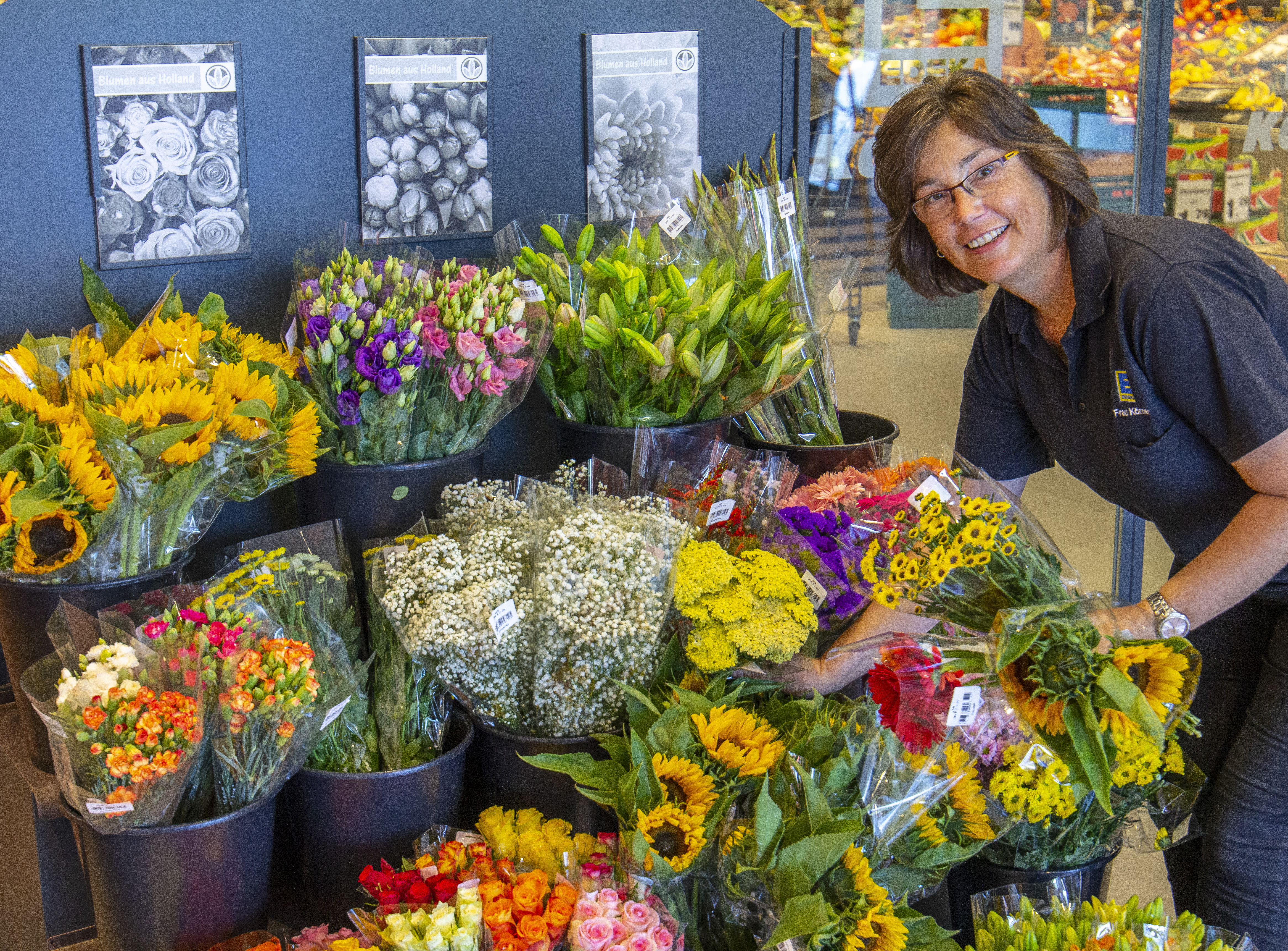 Die Blumenauslage eines EDEKA-Markts. Eine lächelnde Mitarbeiterin steht daneben, weitere Blumensträuße in den Händen.