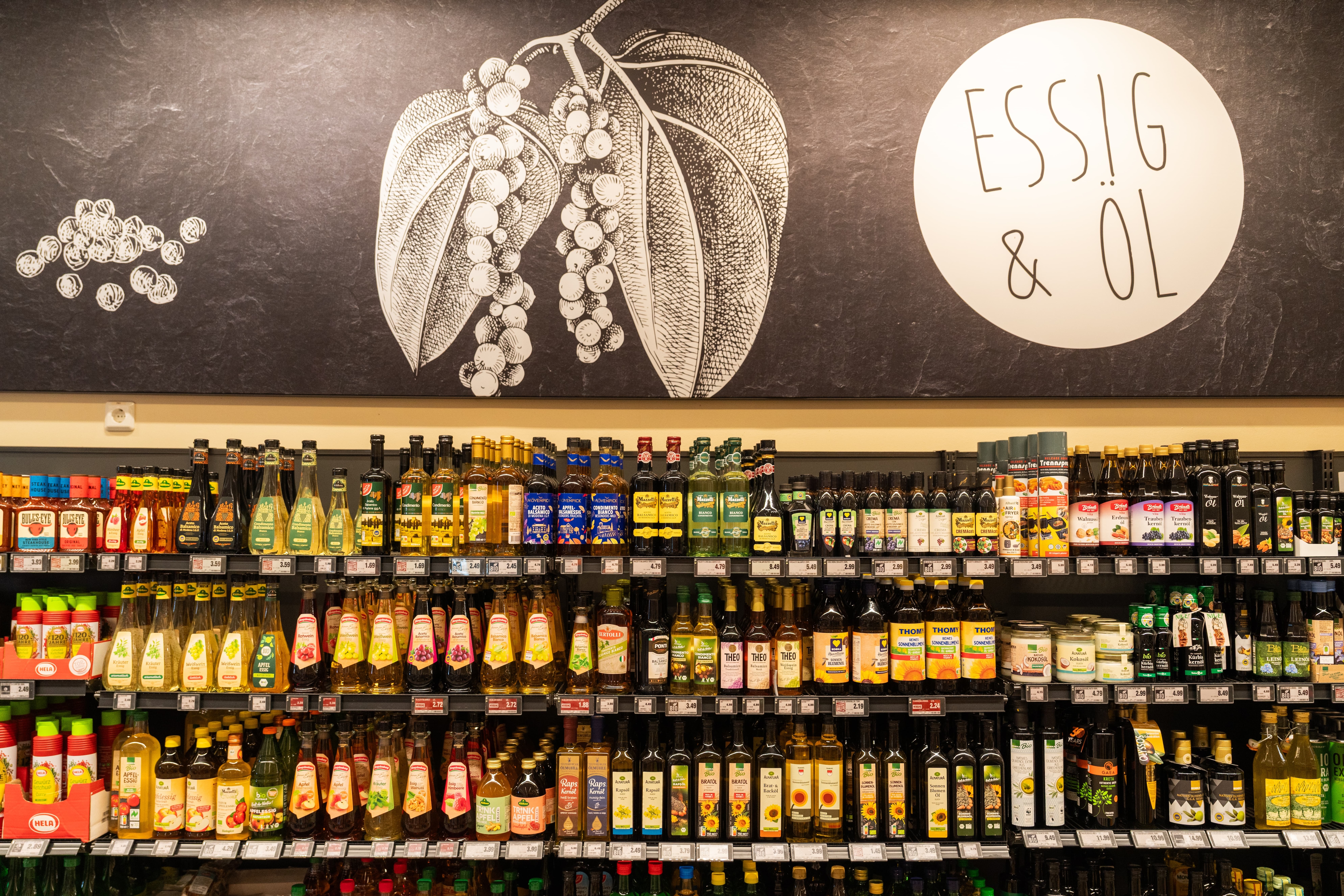Regalreihe mit verschiedenen Essig & Ölsorten. Darüber eine große schwarz/weiß Wandgestaltung passend dazu.