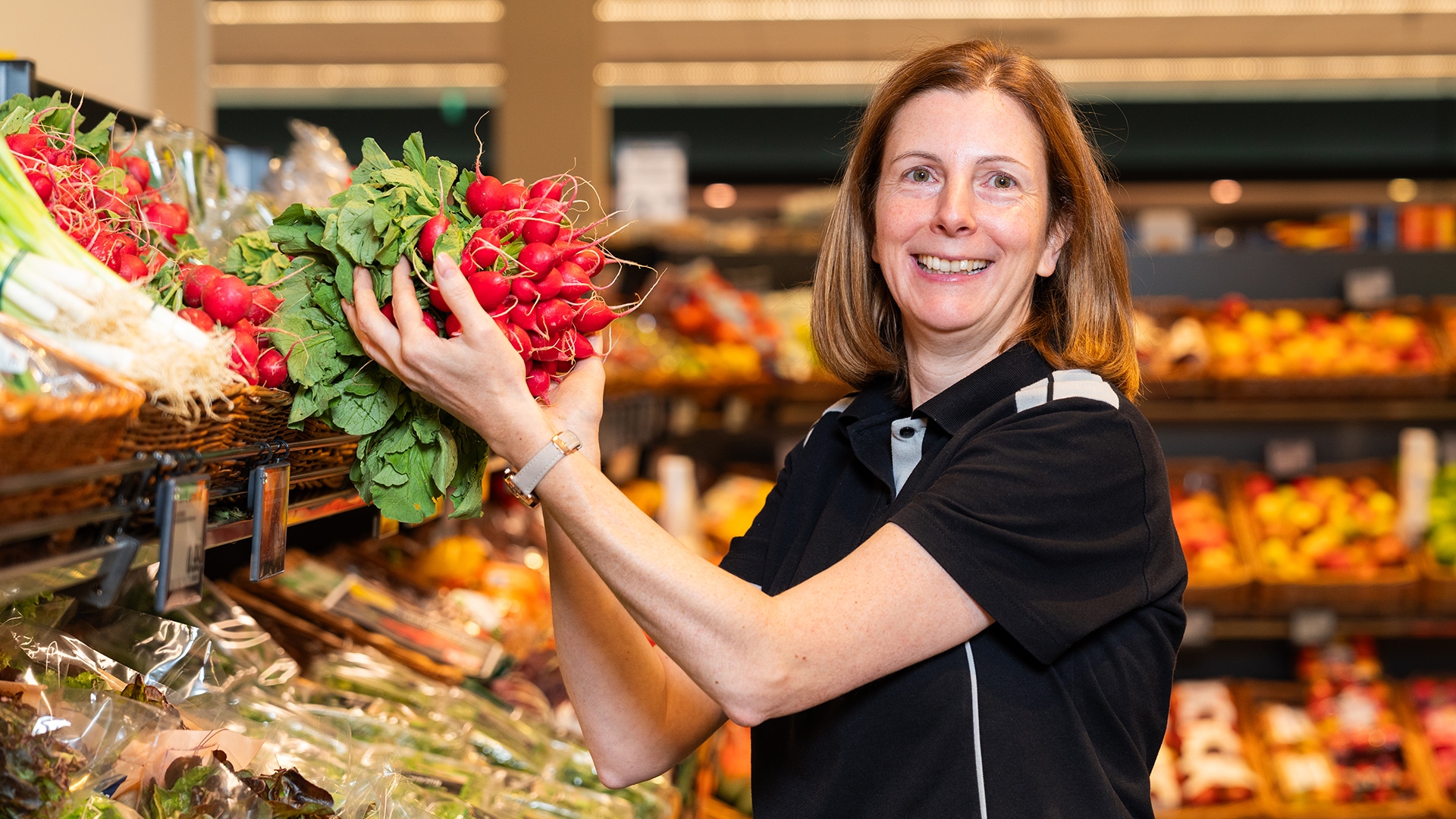 Mitarbeiterin steht mit einem Bund Radieschen in der Hand in der Gemüseabteilung und lächelt in die Kamera.