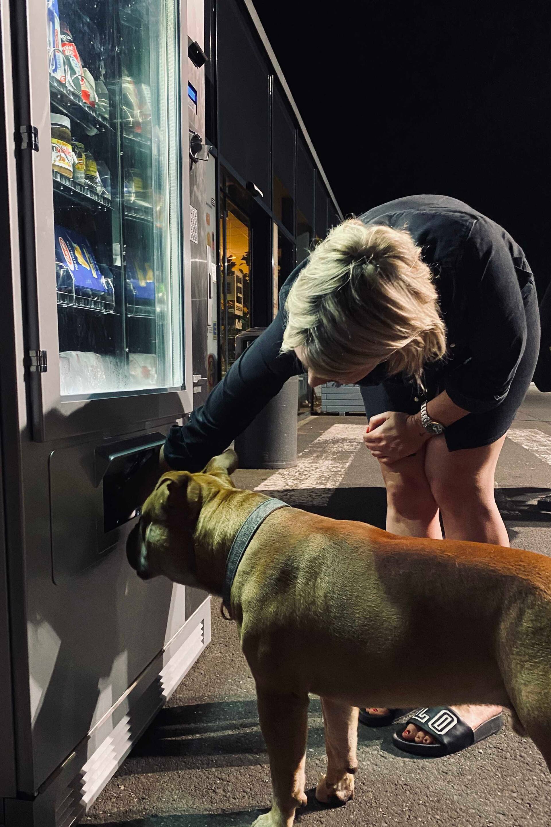 Eine Frau und ihr Hund stehen nachts an einem Snackautomaten. Die Frau nimmt etwas aus einem Snackautomaten.