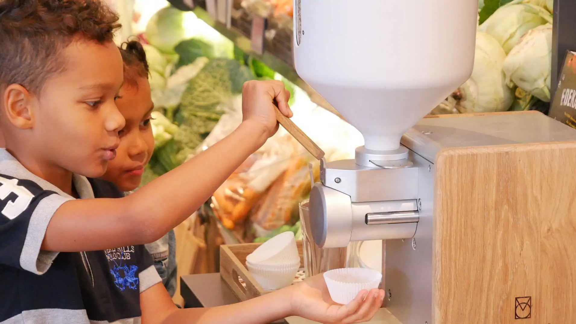Zwei Kinder stehen an einem Spender für Nusscreme. Das eine Kind hält eine kleine Form unter den Spender um sich etwas abzufüllen.