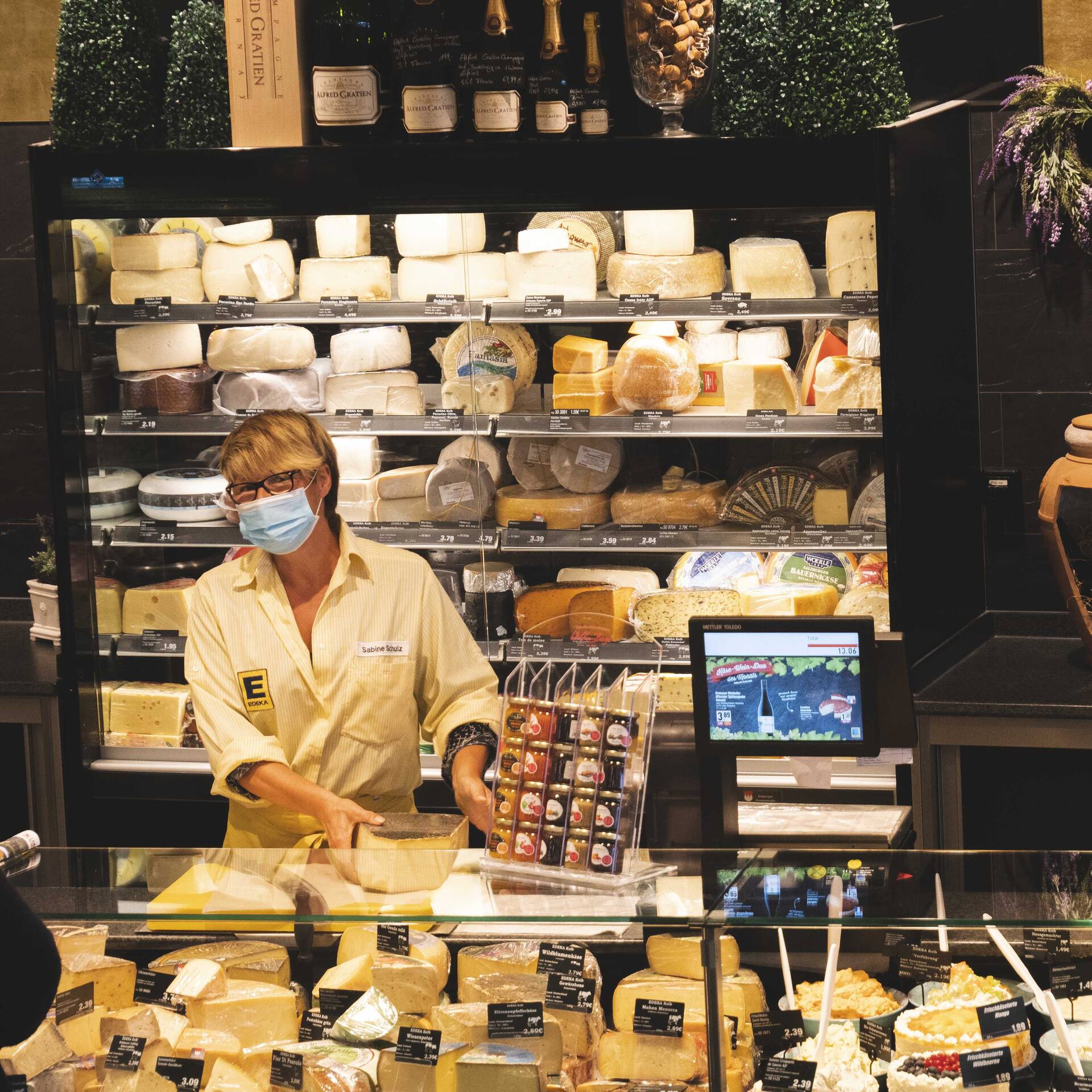 Eine Mitarbeiterin mit Maske steht hinter einer Käsetheke. Im Hintergrund ist ein Kühlregal mit Käse zu sehen.