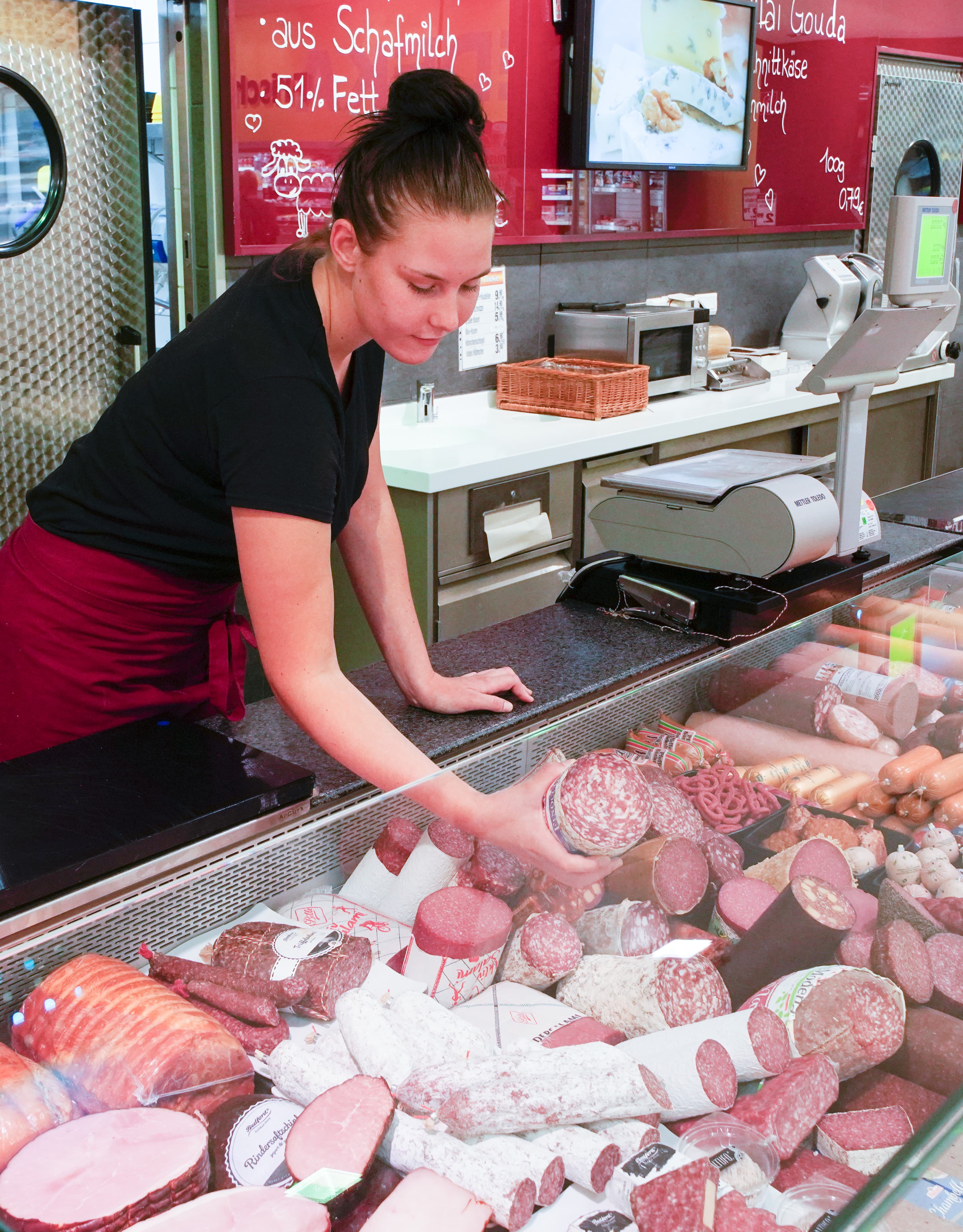 An der Bedientheke eines EDEKA-Markts nimmt eine Mitarbeiterin eine Wurst aus der Auslage.