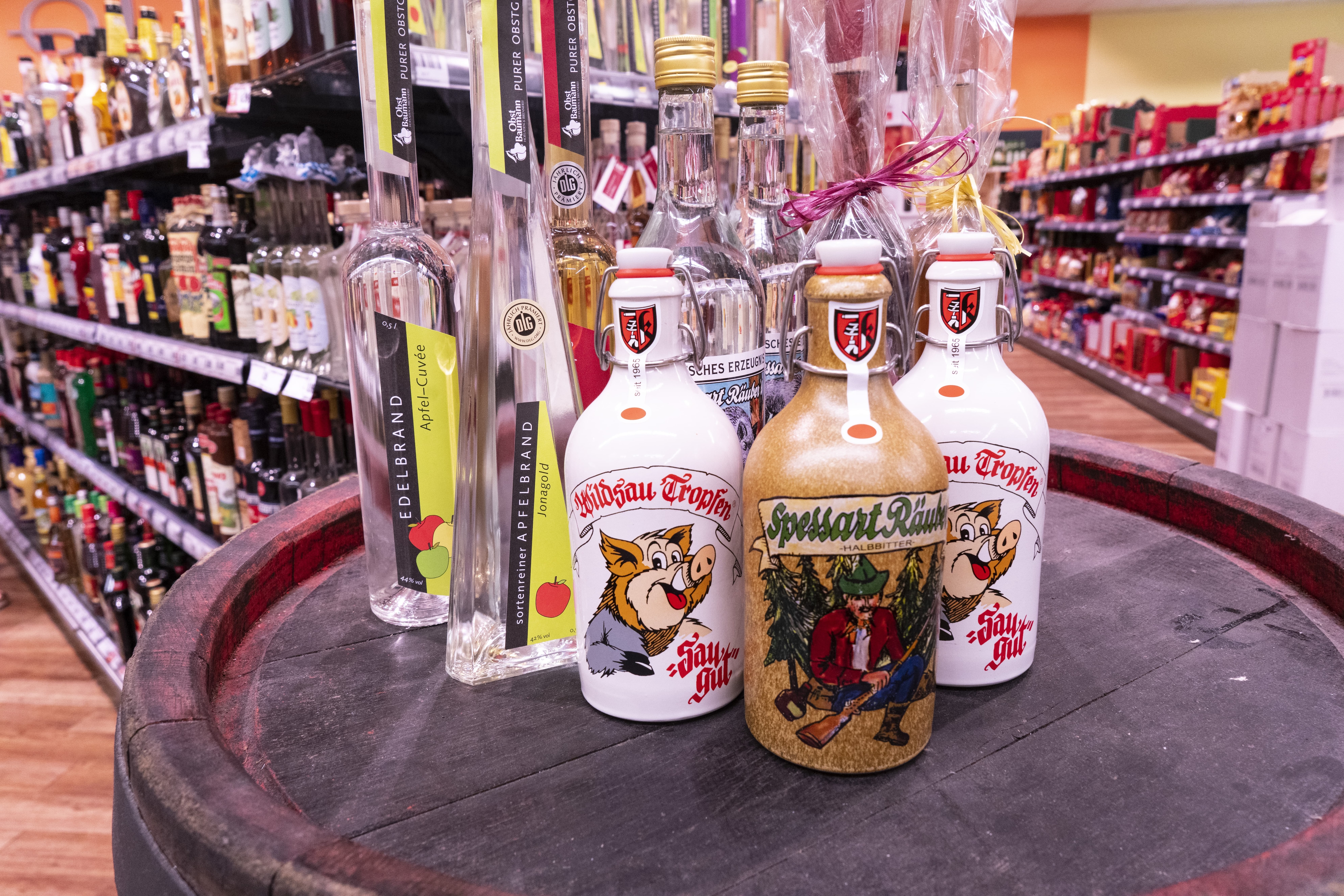 Mehrere alkoholische Getränke aus der Region, angeordnet auf einem Fass, im Hintergrund die Alkoholabteilung des EDEKA-Markts.