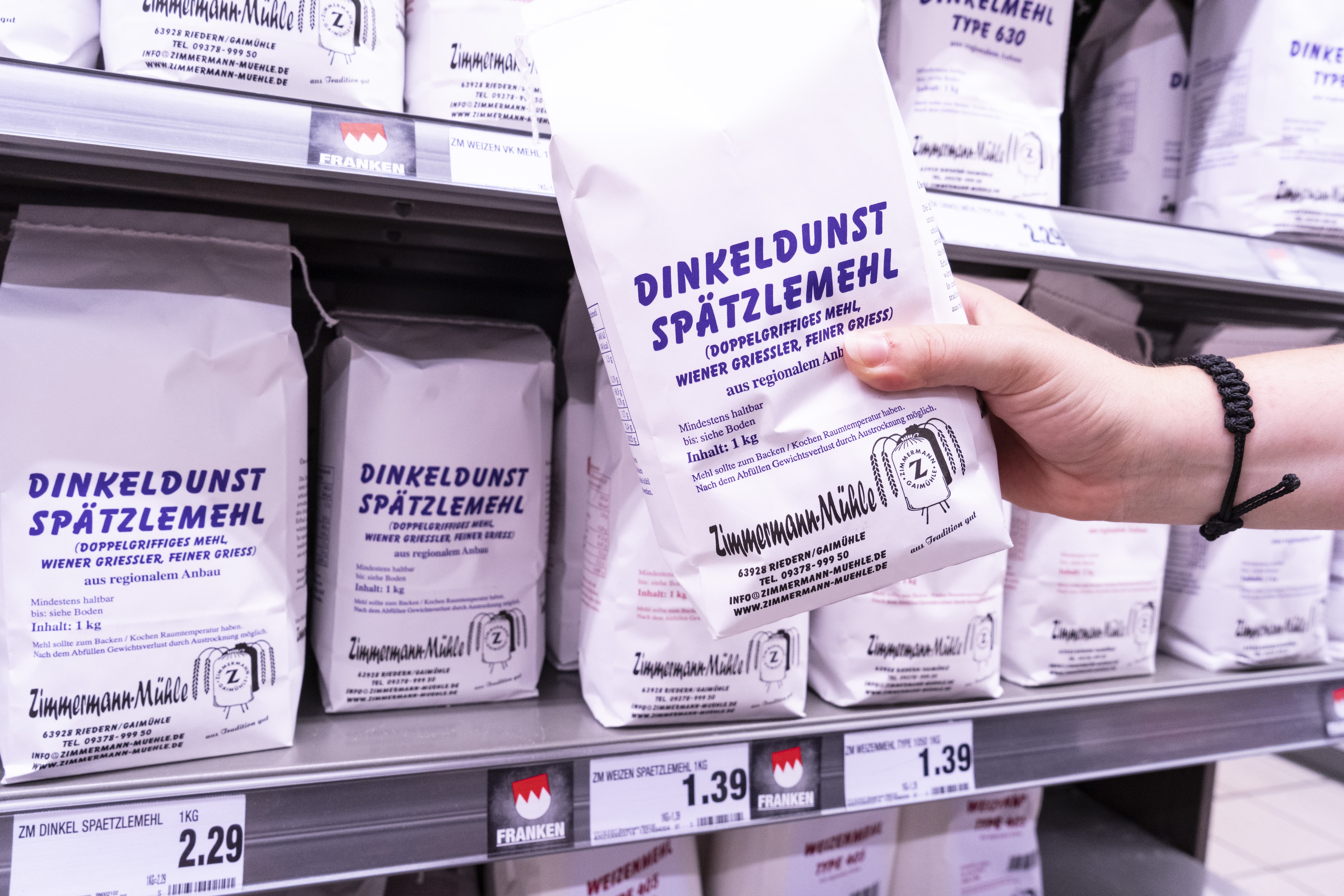 Eine Hand hält eine Papiertüte mit „Dinkeldunst Spätzlemehl“ eines regionalen Herstellers hoch, im Hintergrund Regale mit demselben Produkt.