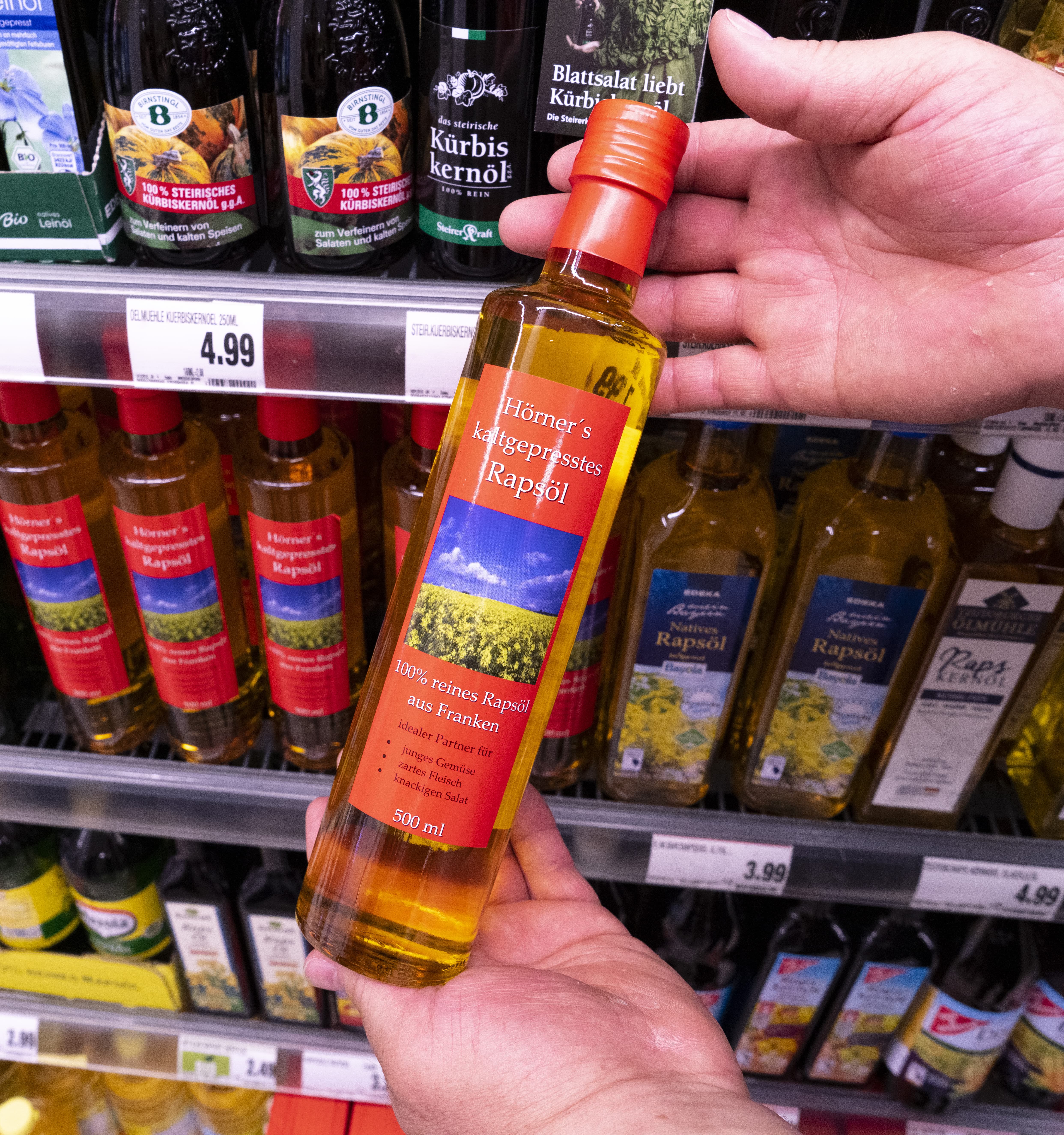 Zwei Hände halten eine Flasche Rapsöl eines regionalen Produzenten hoch, im Hintergrund Regale mit weiteren Ölflaschen.