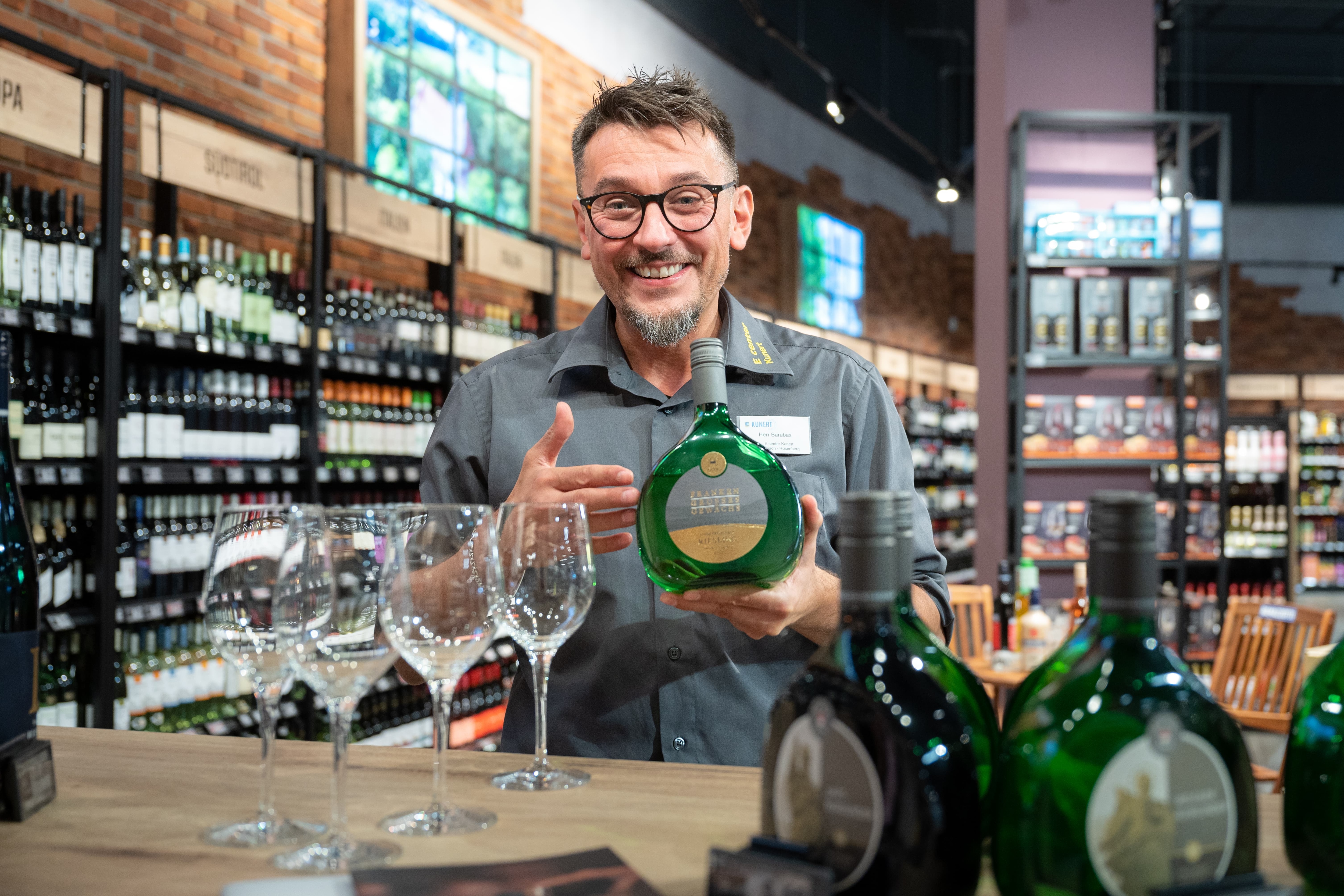 Ein lächelnder Mitarbeiter steht an einem Tisch mit Weingläsern und hält eine Flasche Wein in den Händen.