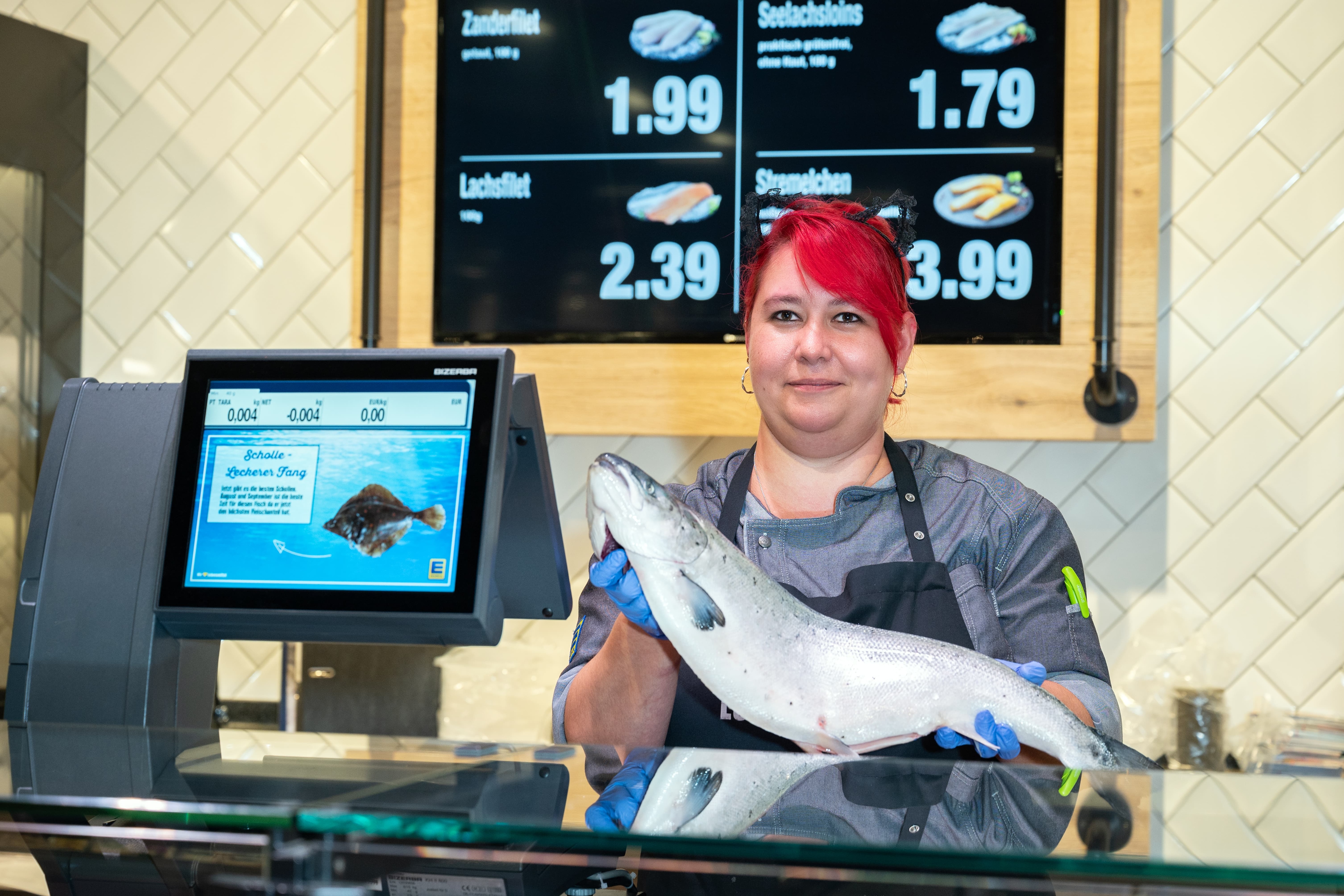 Eine lächelnde Edeka-Mitarbeiter steht hinter einer Theke und hält einen Fisch in den Händen.