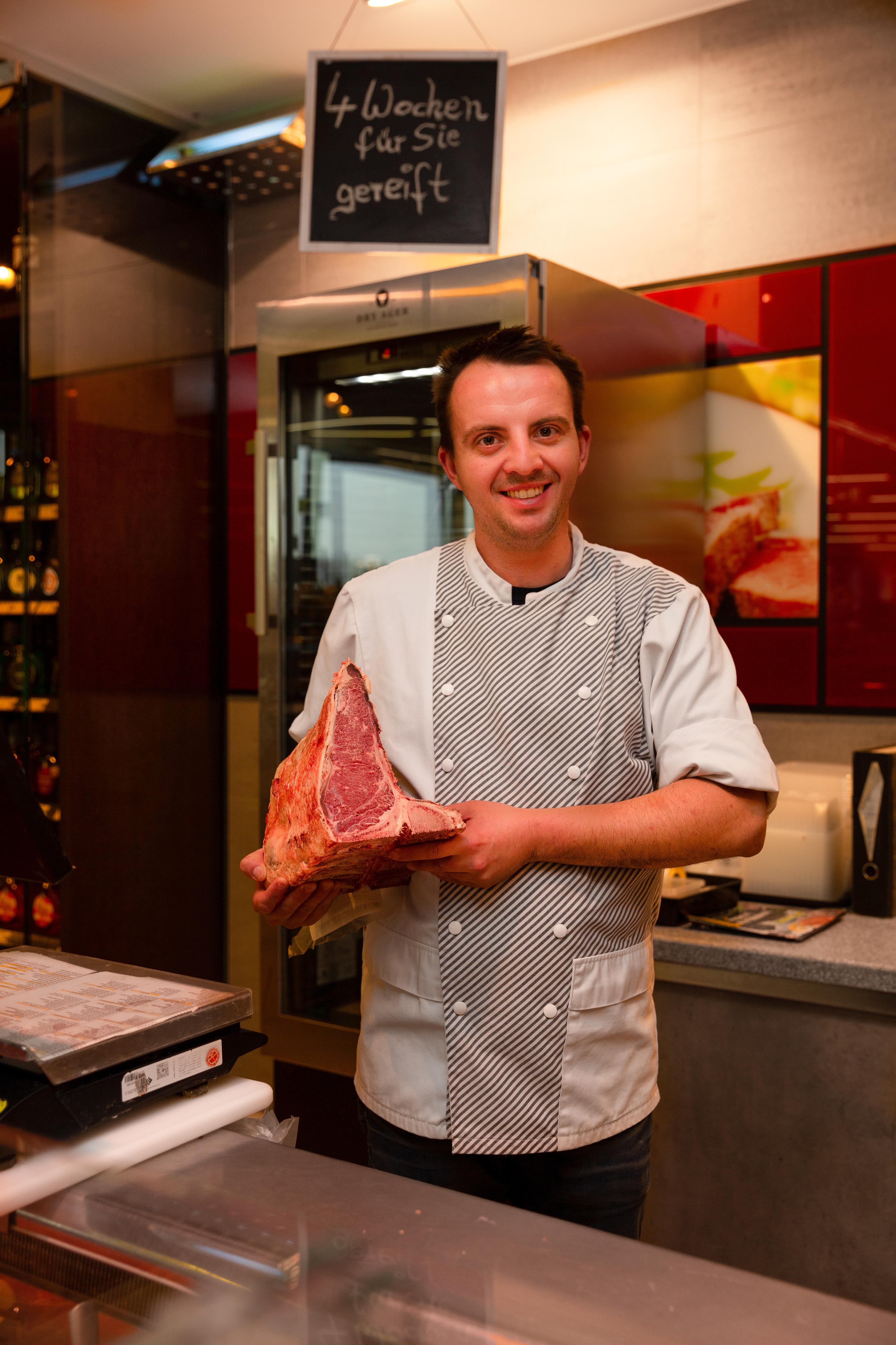 Ein lächelnder Edeka-Mitarbeiter steht hinter der Fleischtheke und hält ein großes Stück Fleisch in den Händen.