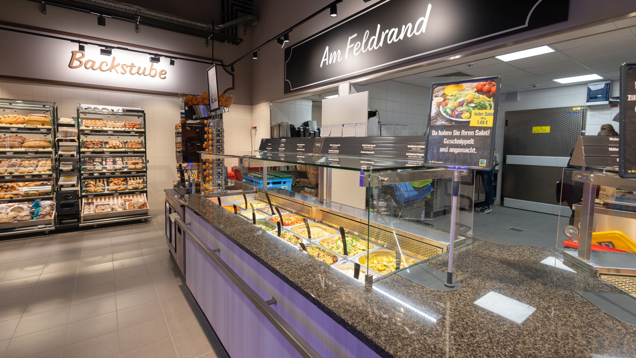 Auf der rechten Seite befindet sich die Frische-Salatbar mit dem Abteilungsschild "Am Feldrand". Geht man an der Salatbar vorbei, erreicht man die Regale der "Backstube" mit verschiedenen Brot- und Brötchensorten