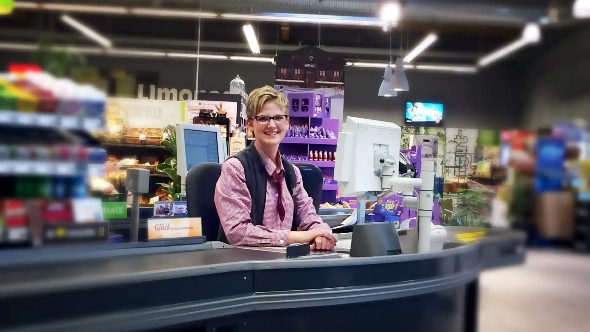 Eine lächelnde Mitarbeiterin sitzt an der Kasse eines EDEKA-Marktes.