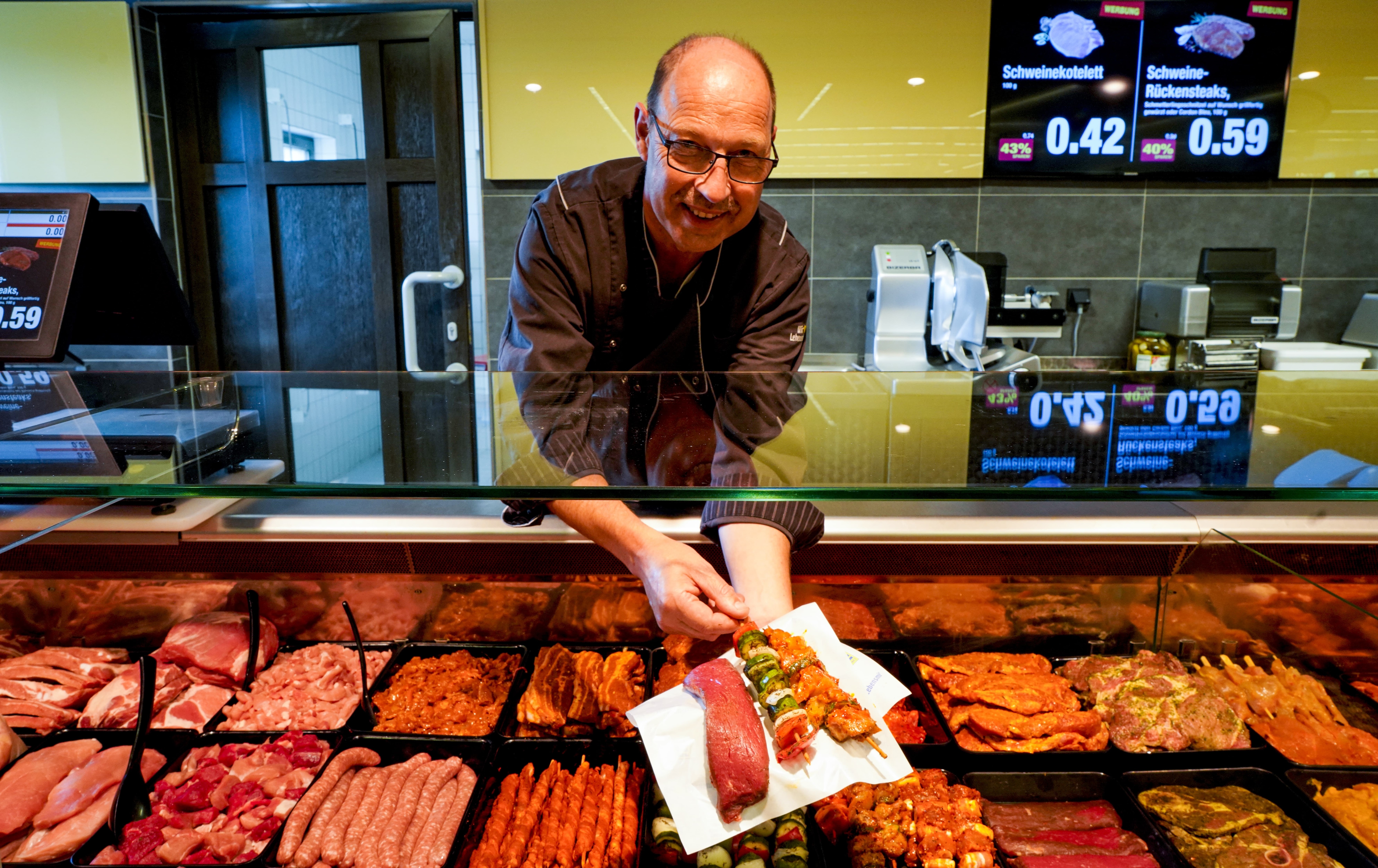 Ein Mitarbeiter an der Fleischtheke eines EDEKA-Markts hält lächelnd einige Fleischspieße in die Kamera.