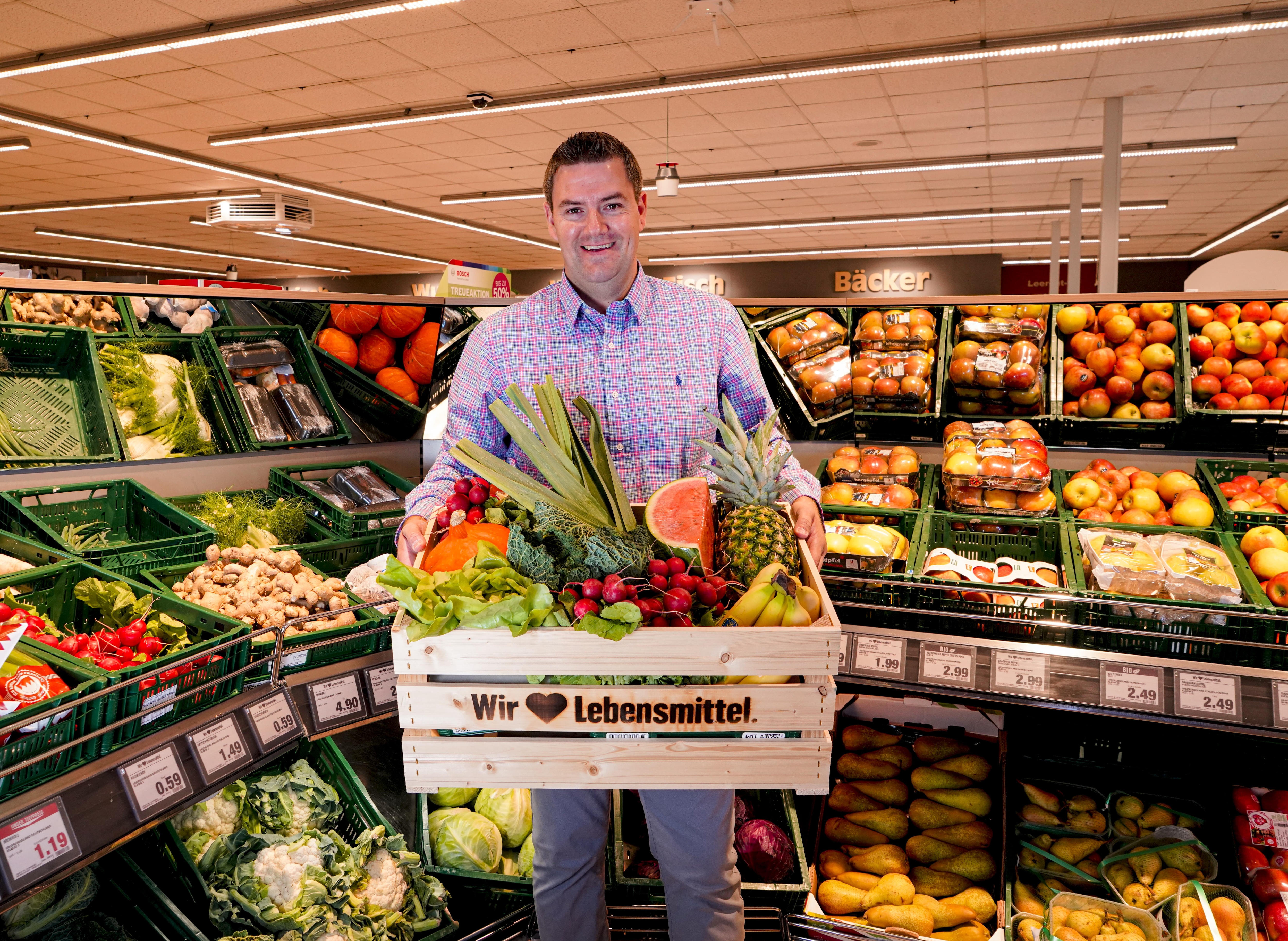 Der Marktleiter, lächelnd mit einer Kiste voller Gemüse im Arm, im Hintergrund die Obst- und Gemüseabteilung.