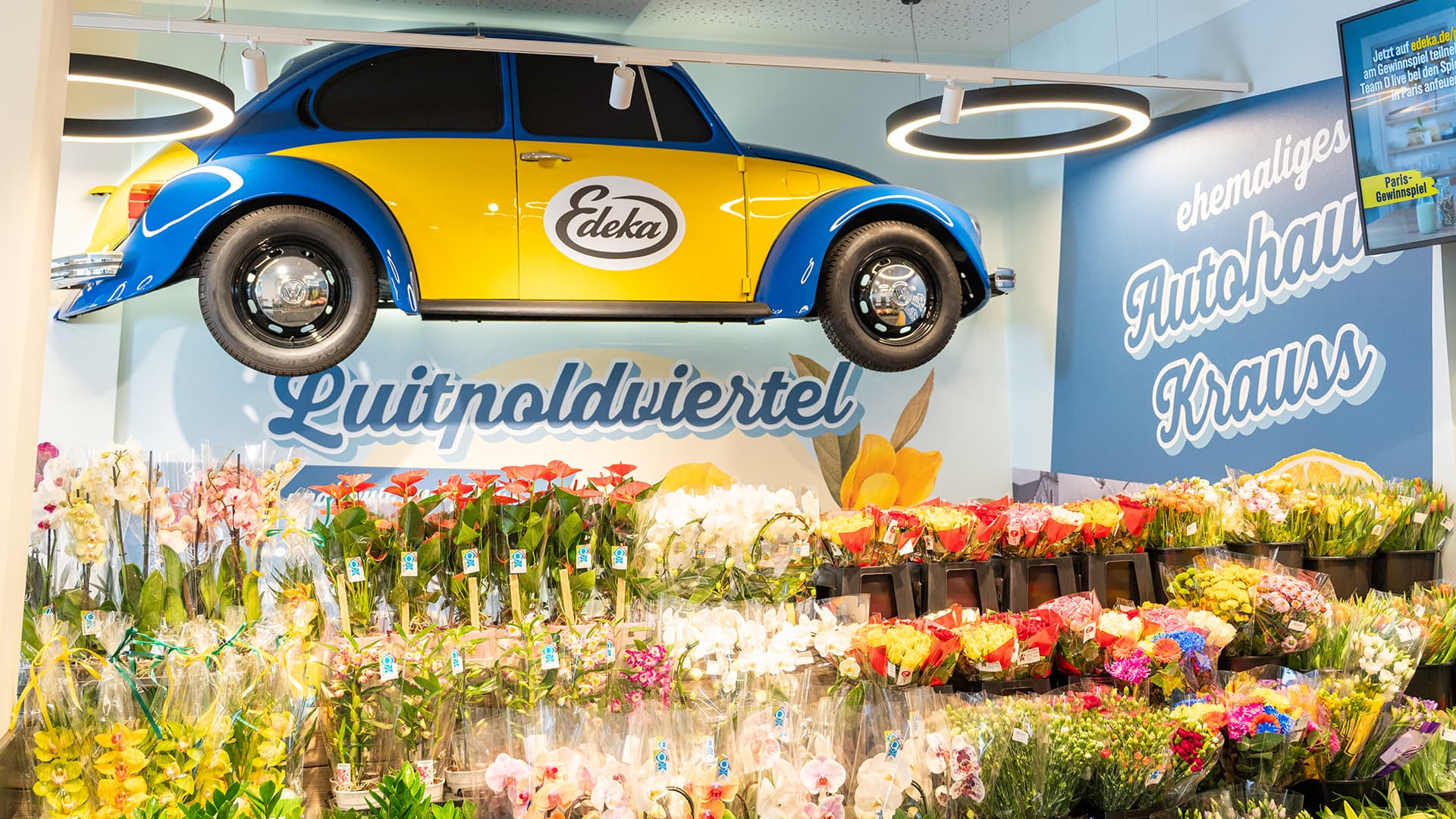 Ein Regal mit Blumen. An der Wand darüber hängt ein blau-gelber VW-Beatle mit einem Edeka-Schriftzug auf der Tür.